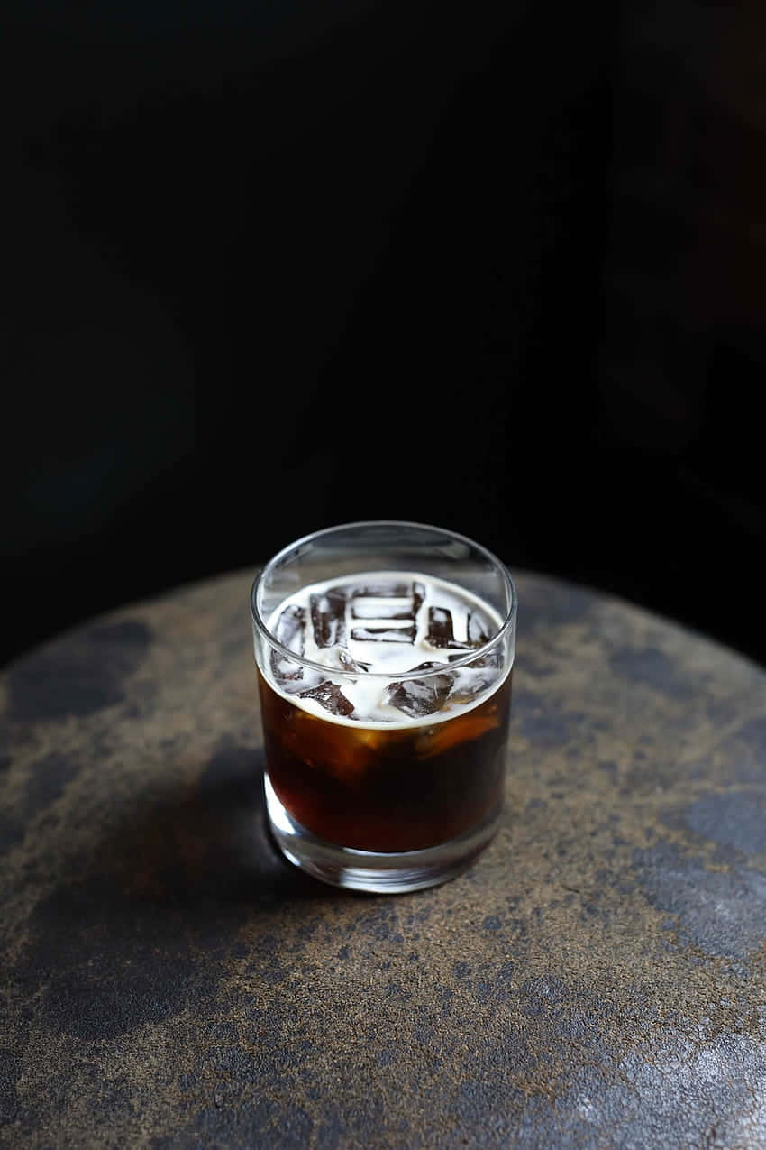 Iced Coffee 桌布 壁紙 — 一杯深琥珀色的雞尾酒加冰塊，放在透明玻璃杯中，置於有紋理的石面上，背景為黑色。