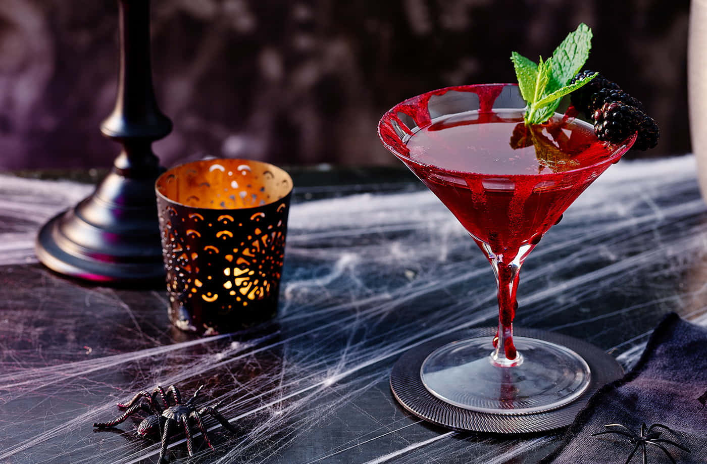 Halloween Cocktails 桌布 壁紙 — 一杯深紅色雞尾酒，飾以薄荷和黑莓，放在鋪滿蜘蛛網的桌子上，旁邊有一根閃爍的蠟燭和塑膠蜘蛛。