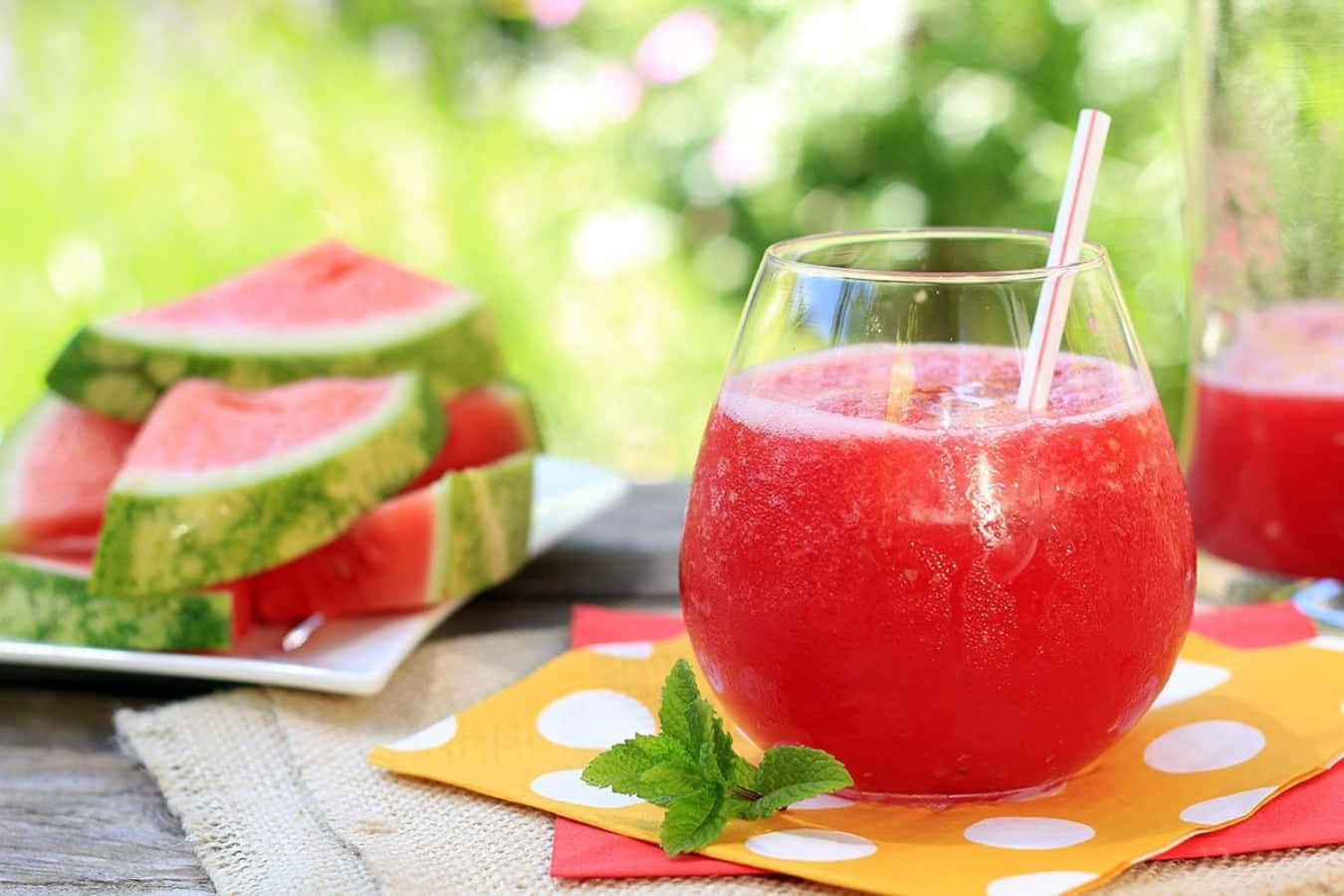 Watermelon 桌布 壁紙 — 一杯清涼的西瓜冰沙搭配薄荷裝飾，放在圓點餐巾上，旁邊是新鮮西瓜片，在戶外。