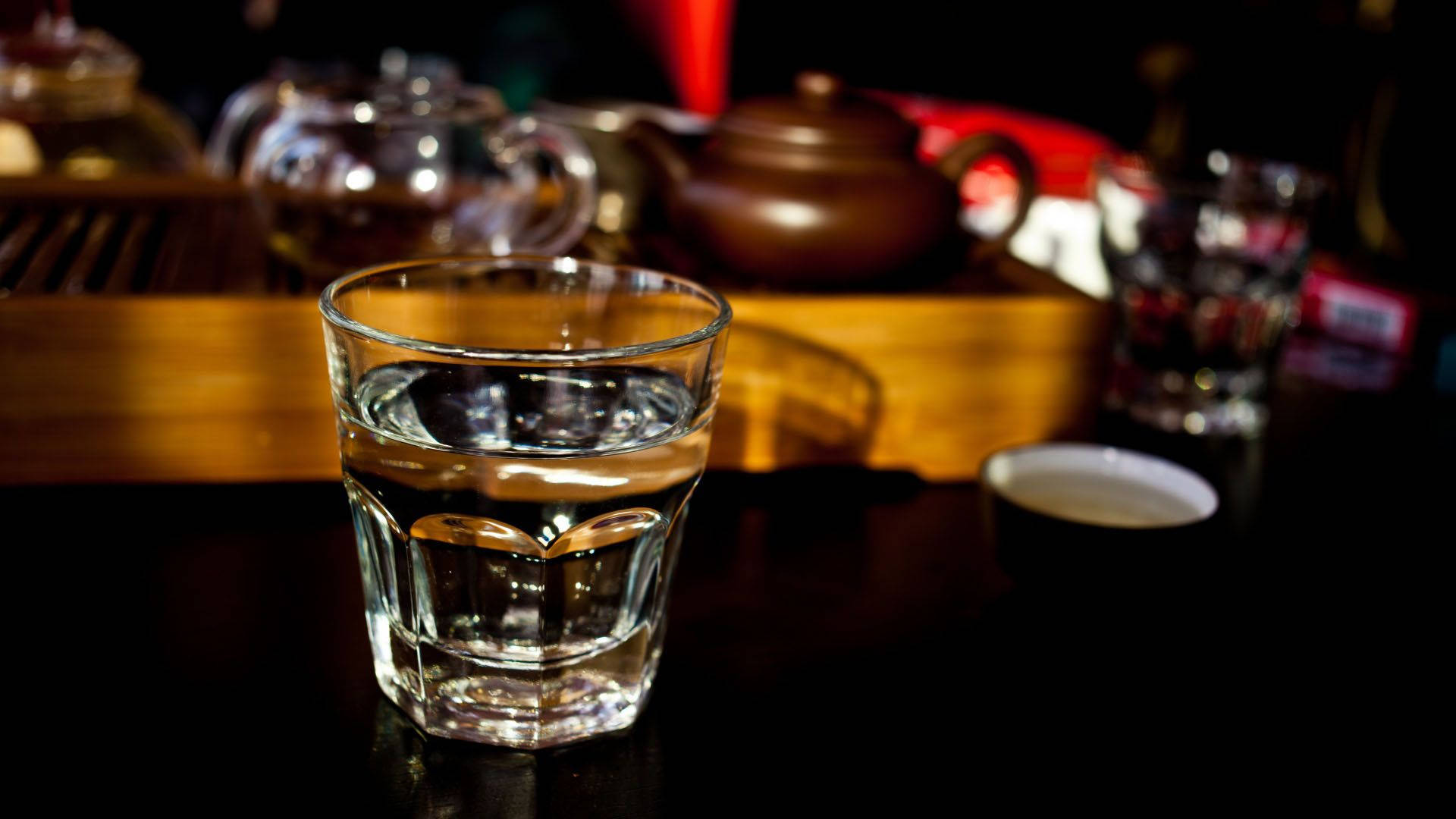 Drink 桌布 壁紙 — 一杯清澈的水在焦點中，放在一張深色桌子上，背景有溫暖的燈光下模糊的茶具。