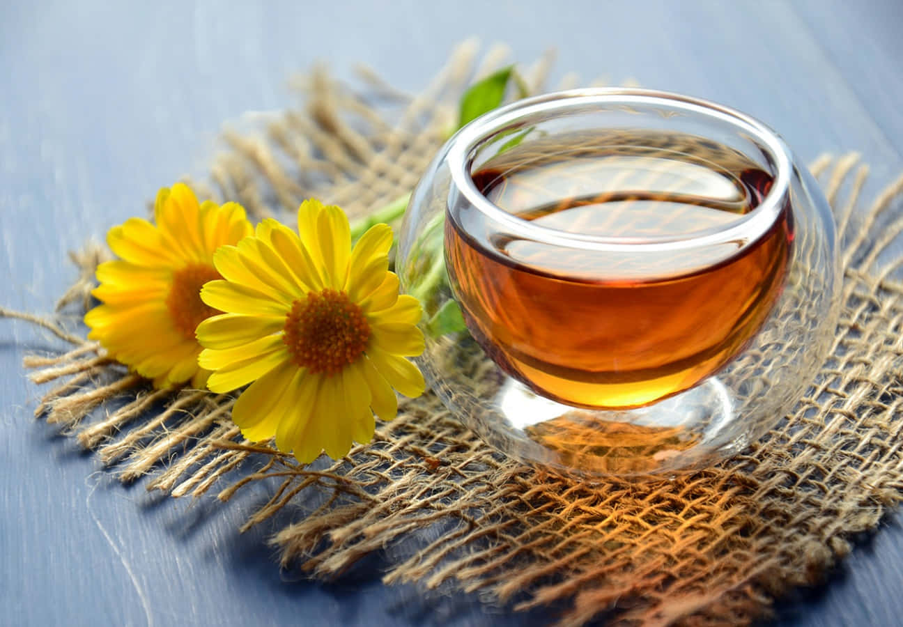 tea 桌布 壁紙 — 一杯清澈的琥珀色茶放在藍色木質表面上，旁邊是樸素的黃麻墊上的鮮黃色雛菊。