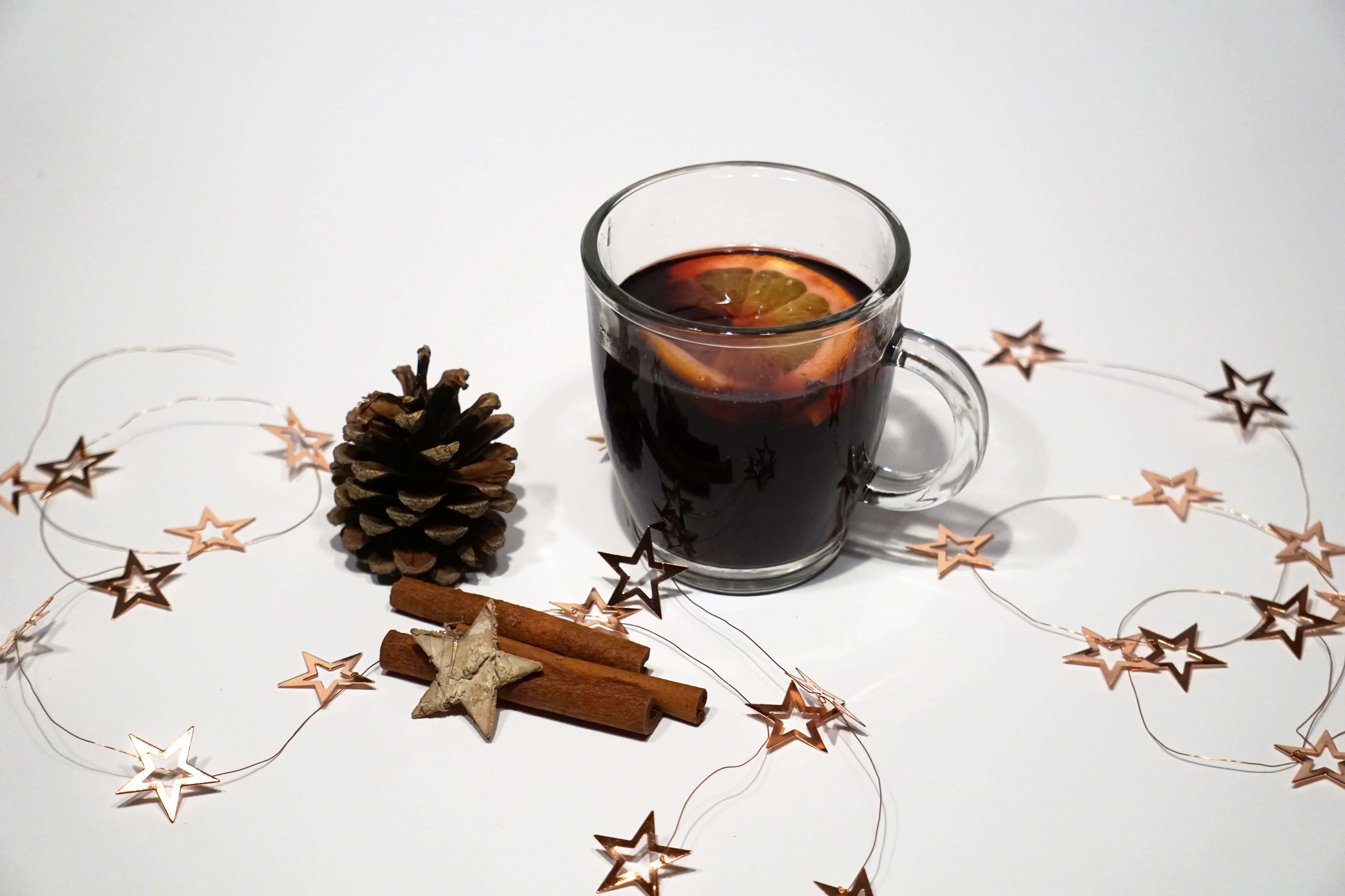Mulled Wine 桌布 壁紙 — 一杯清澈的香料熱紅酒配橙片，旁邊放著松果、肉桂棒和銅星花環，置於白色表面上。