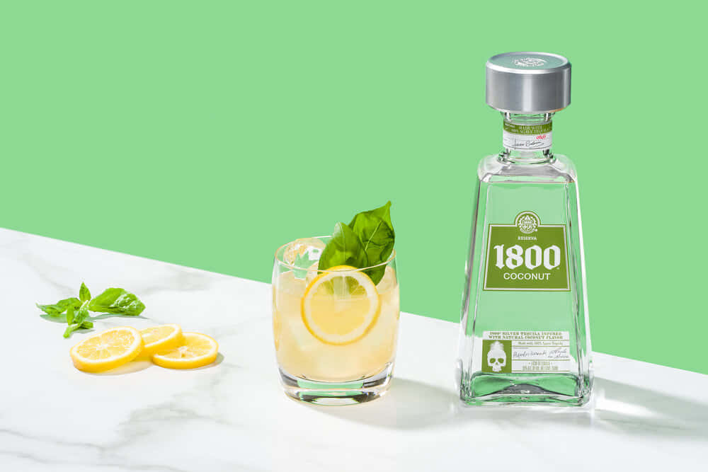 alcohol 桌布 壁紙 — 一杯清爽的1800椰子龍舌蘭雞尾酒，飾以檸檬和羅勒，旁邊放著酒瓶，置於白色大理石表面上，背景是鮮豔的綠色。