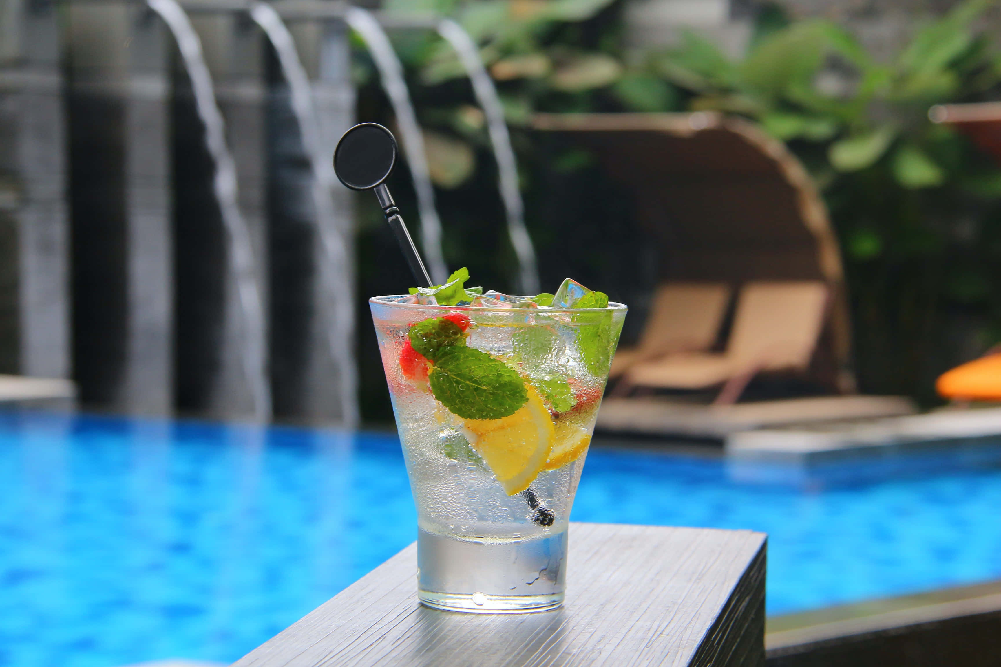 Summer Drinks 桌布 壁紙 — 一杯清爽的柑橘雞尾酒，配上薄荷和冰塊，放在木製邊緣上，背景是層疊的泳池和躺椅。