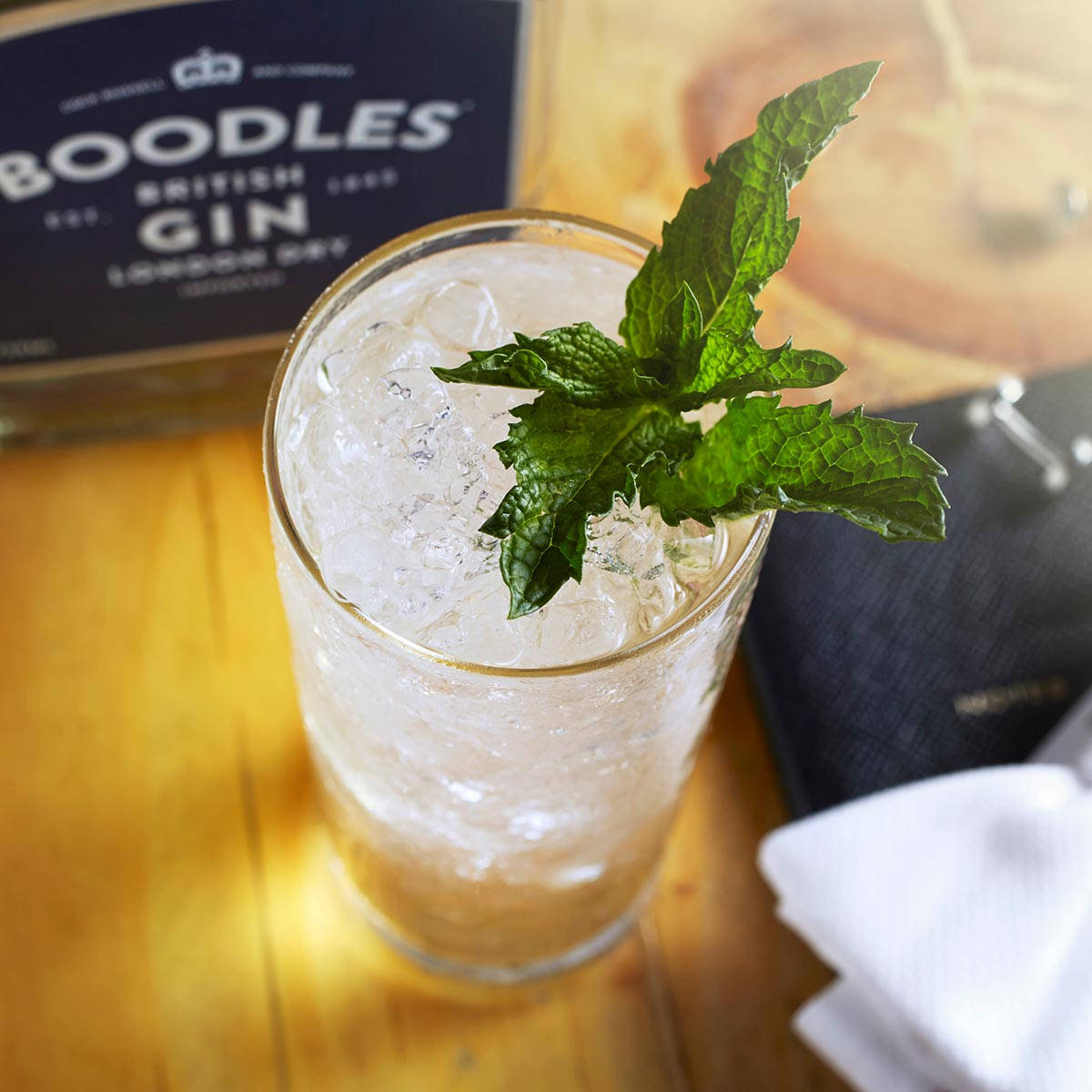 cocktail 桌布 壁紙 — 一杯清爽的琴酒雞尾酒，配上薄荷和冰塊，放在木桌上，旁邊有一個Boodles琴酒盒和一條深色餐巾。