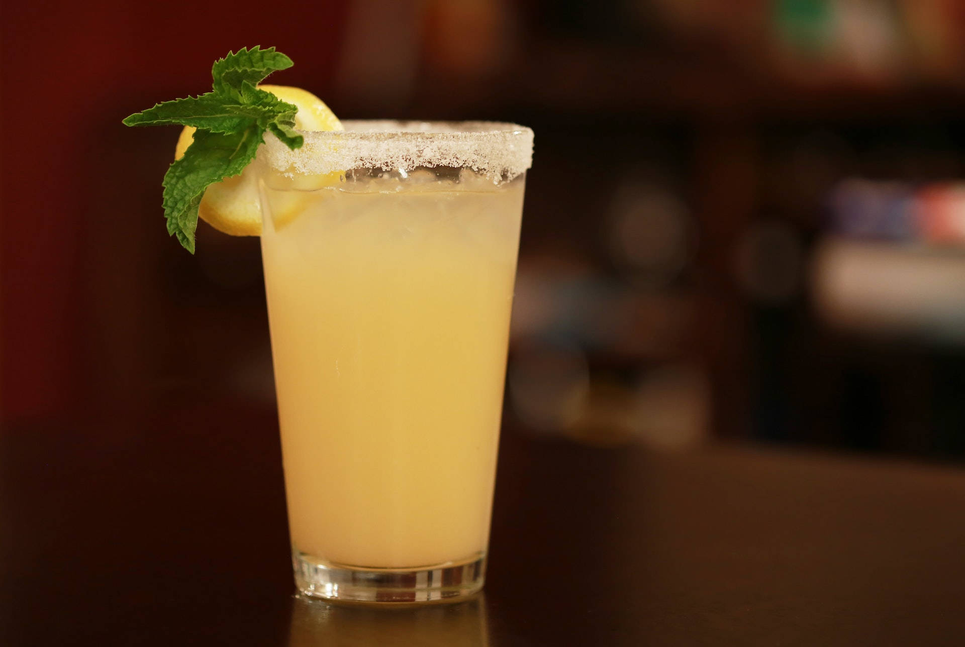 alcohol 桌布 壁紙 — 一杯清爽的瑪格麗特雞尾酒，帶有鹽邊、檸檬角和薄荷裝飾，放在深色吧檯上，背景是模糊的暖色調。