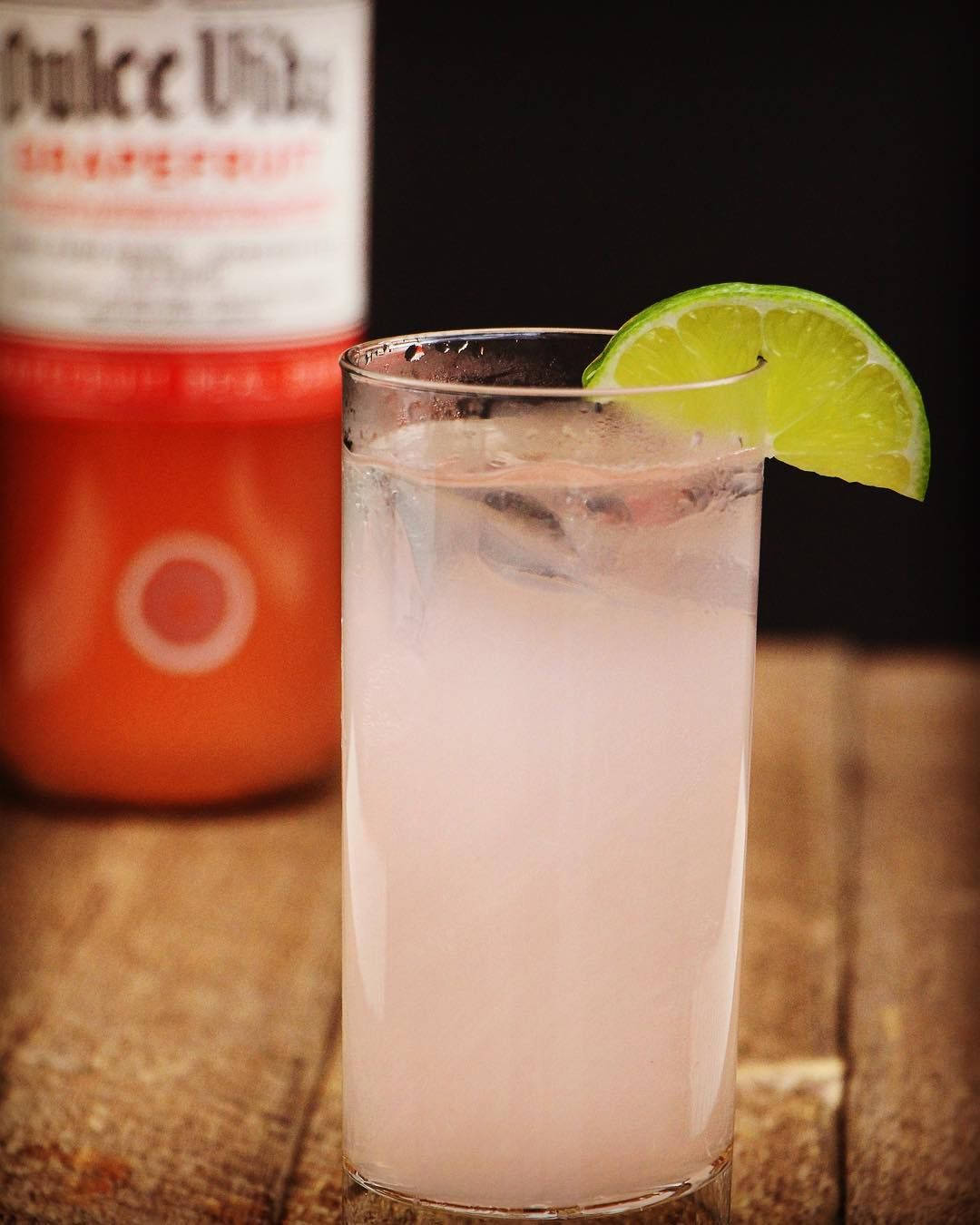 alcohol 桌布 壁紙 — 一杯清爽的粉紅色雞尾酒，加冰和萊姆裝飾，放在玻璃杯中，旁邊有一瓶果汁，置於木質表面上。