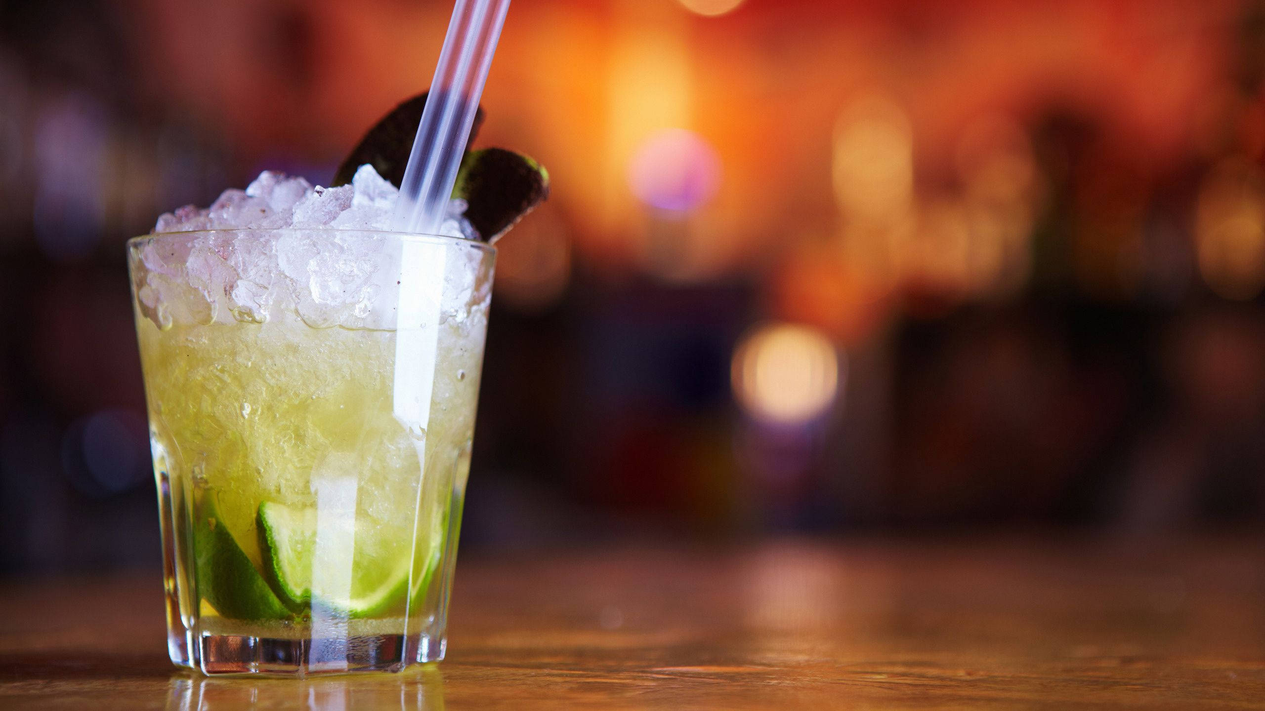 Drink 桌布 壁紙 — 一杯清爽的萊姆雞尾酒，配上碎冰和吸管，放在木製吧檯上，背景是溫暖的散景燈光。