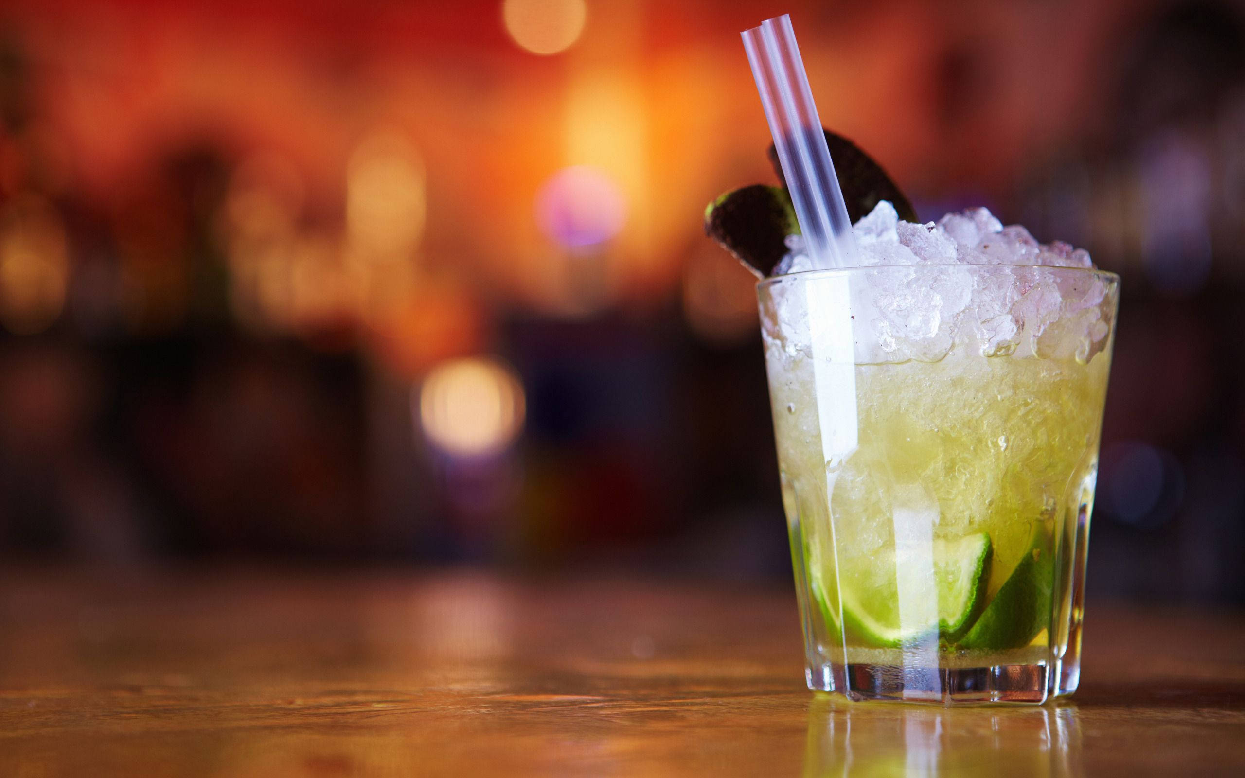 Drink 桌布 壁紙 — 一杯清爽的萊姆雞尾酒，配上碎冰和吸管，放在木製吧檯上，背景是溫暖的散景燈光。