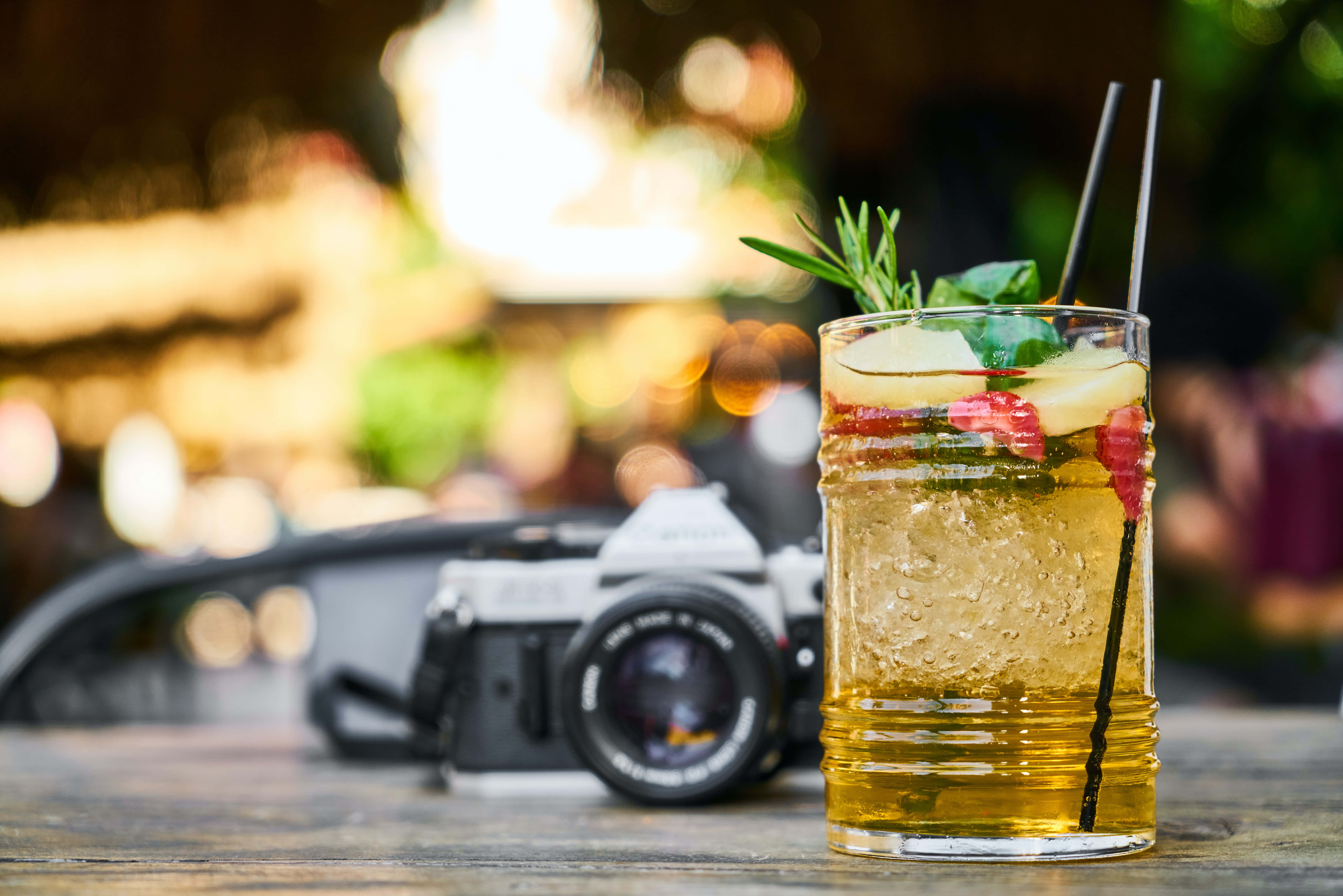Summer Drinks 桌布 壁紙 — 一杯清爽的金色雞尾酒，用迷迭香和水果裝飾，放在一張木桌上，旁邊有一台復古相機，背景是燈光溫暖的戶外酒吧。