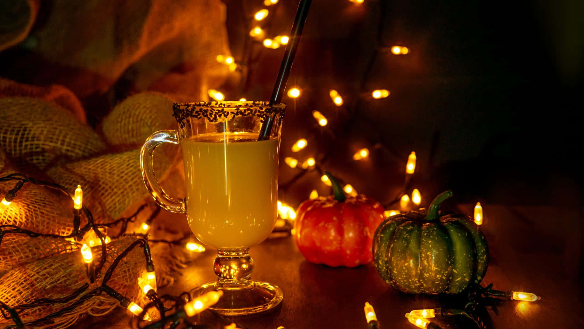 Spooky Cocktails 桌布 壁紙 — 一杯溫暖發光的香料蘋果酒（杯緣沾有糖）放在節日南瓜和閃爍的仙女燈旁，營造出舒適的秋日氛圍。