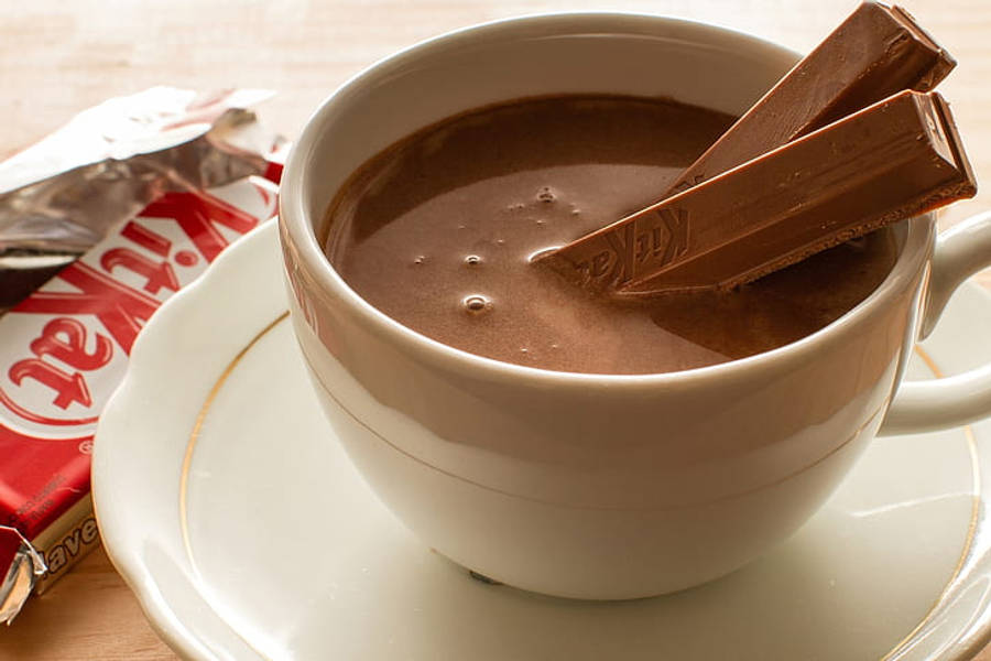 Chocolate 桌布 壁紙 — 一杯濃郁的熱巧克力放在白色陶瓷杯中，兩條 KitKat 浸入其中，置於碟上，旁邊有一條包裝好的 KitKat 在木桌上。