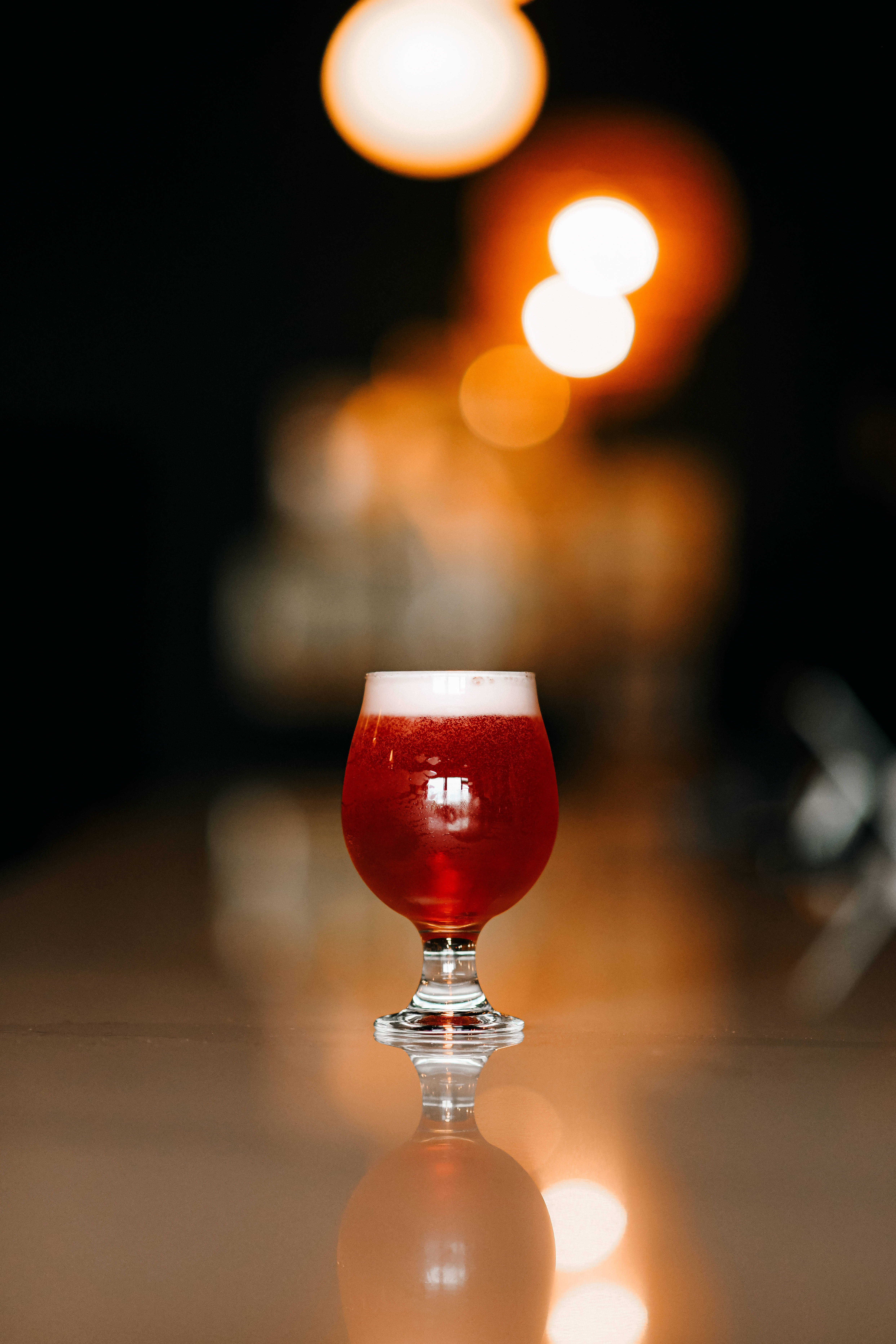 Beer 桌布 壁紙 — 一杯濃郁的紅啤酒，帶有白色泡沫，盛在鬱金香杯中，放在反射的吧檯上，背景是溫暖的散景燈光。