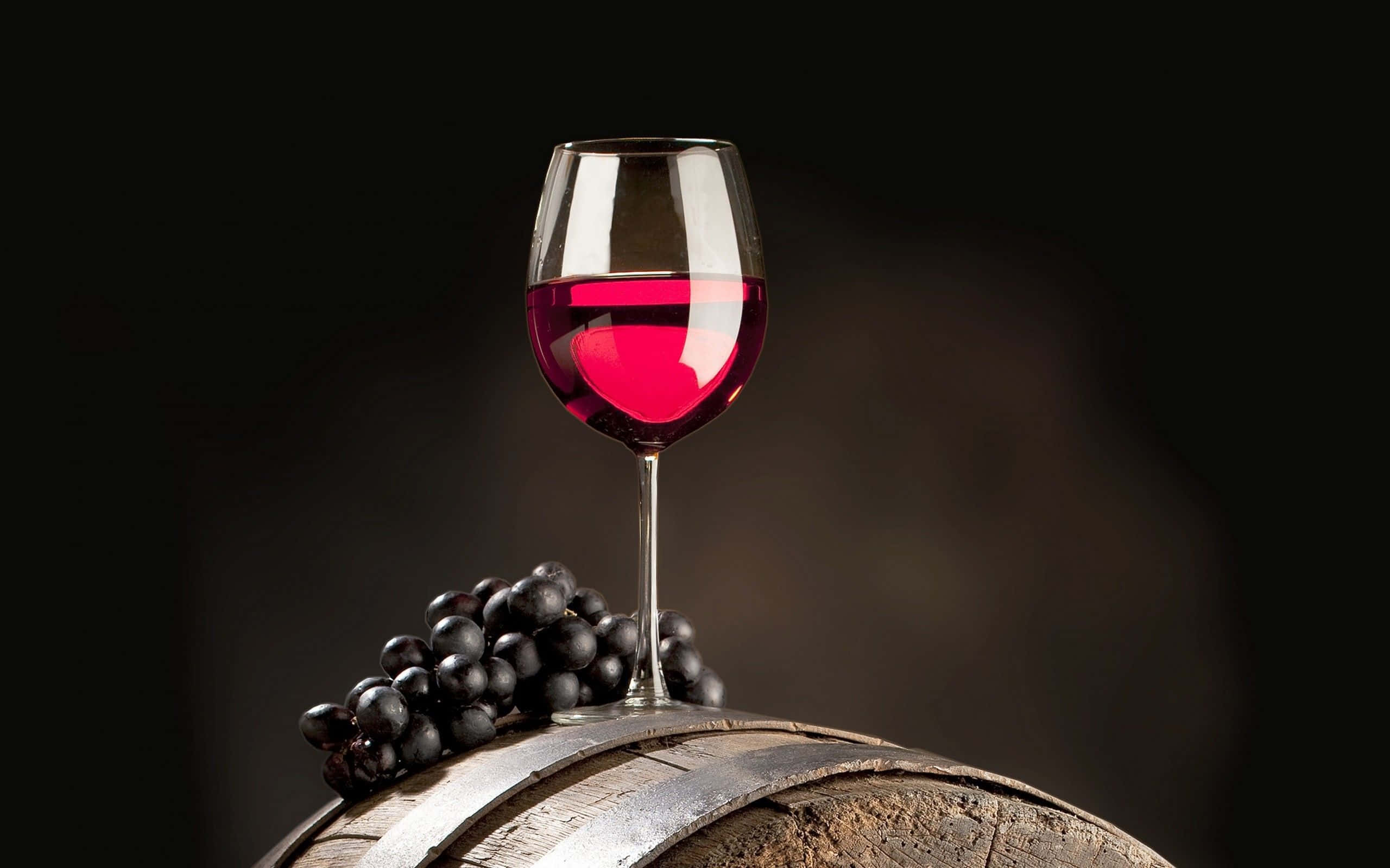 Red Wine Glass 桌布 壁紙 — 一杯濃郁的紅酒優雅地放在一個風化的木桶上，旁邊是一串深色葡萄，背景是深邃的黑色。