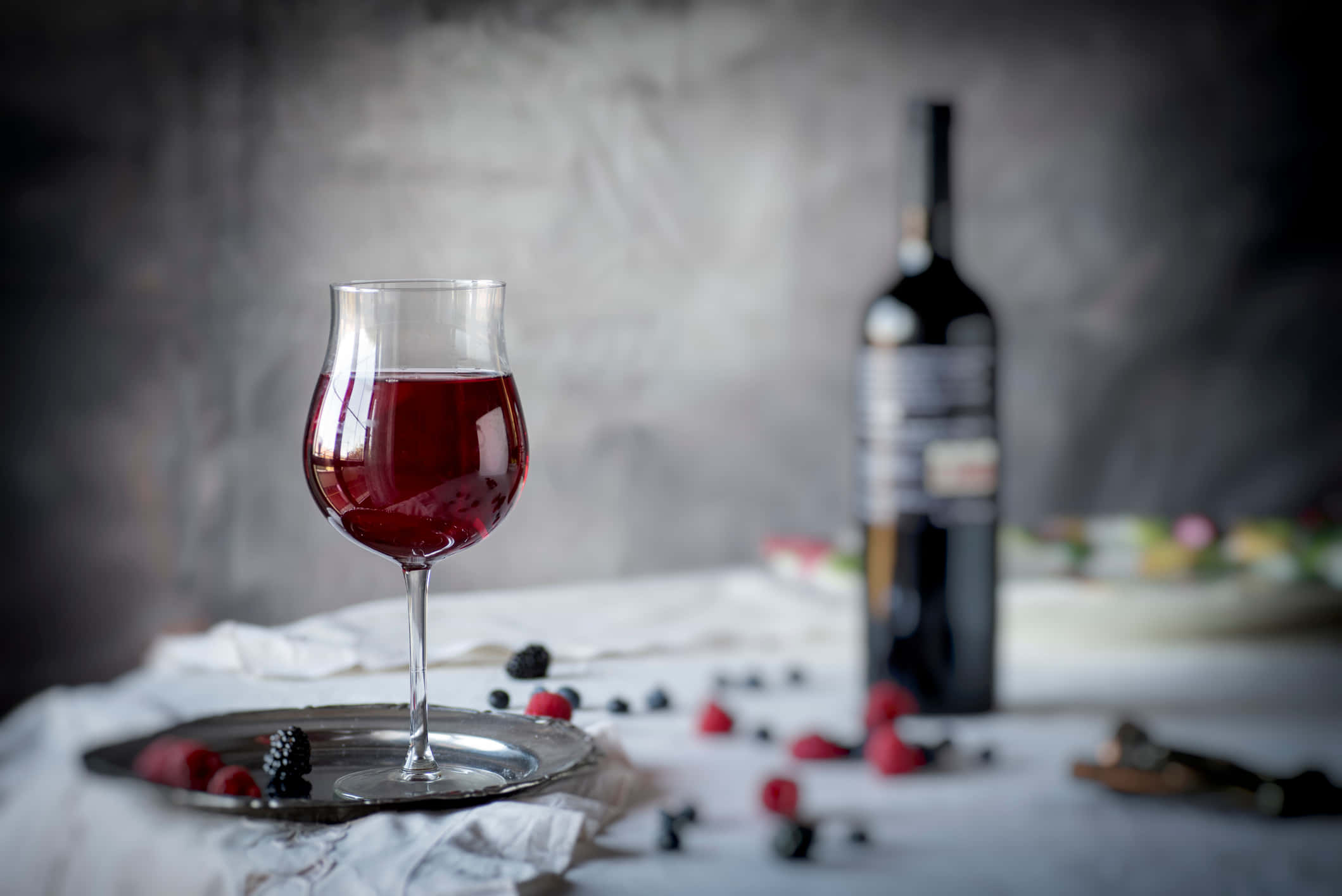 Blackberry Wine 桌布 壁紙 — 一杯濃郁的紅酒放在銀托盤上，旁邊有一瓶酒和散落的莓果，置於有紋理的白色布料上，背景為柔和的灰色。