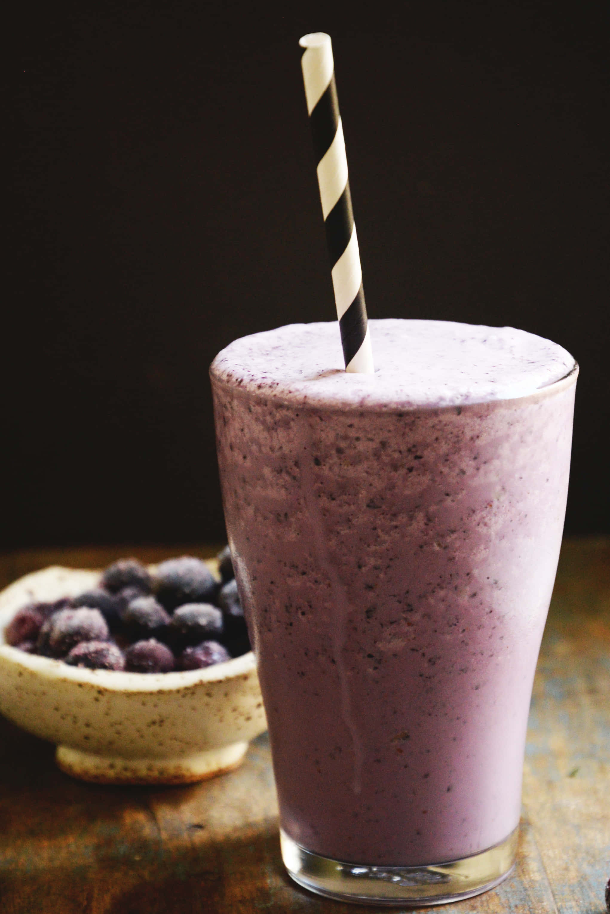 Blueberry Smoothie 桌布 壁紙 — 一杯濃郁的紫色藍莓冰沙，裝在高身玻璃杯中，插著一條條紋吸管，旁邊放著一碗冷凍藍莓，置於鄉村風木桌上。