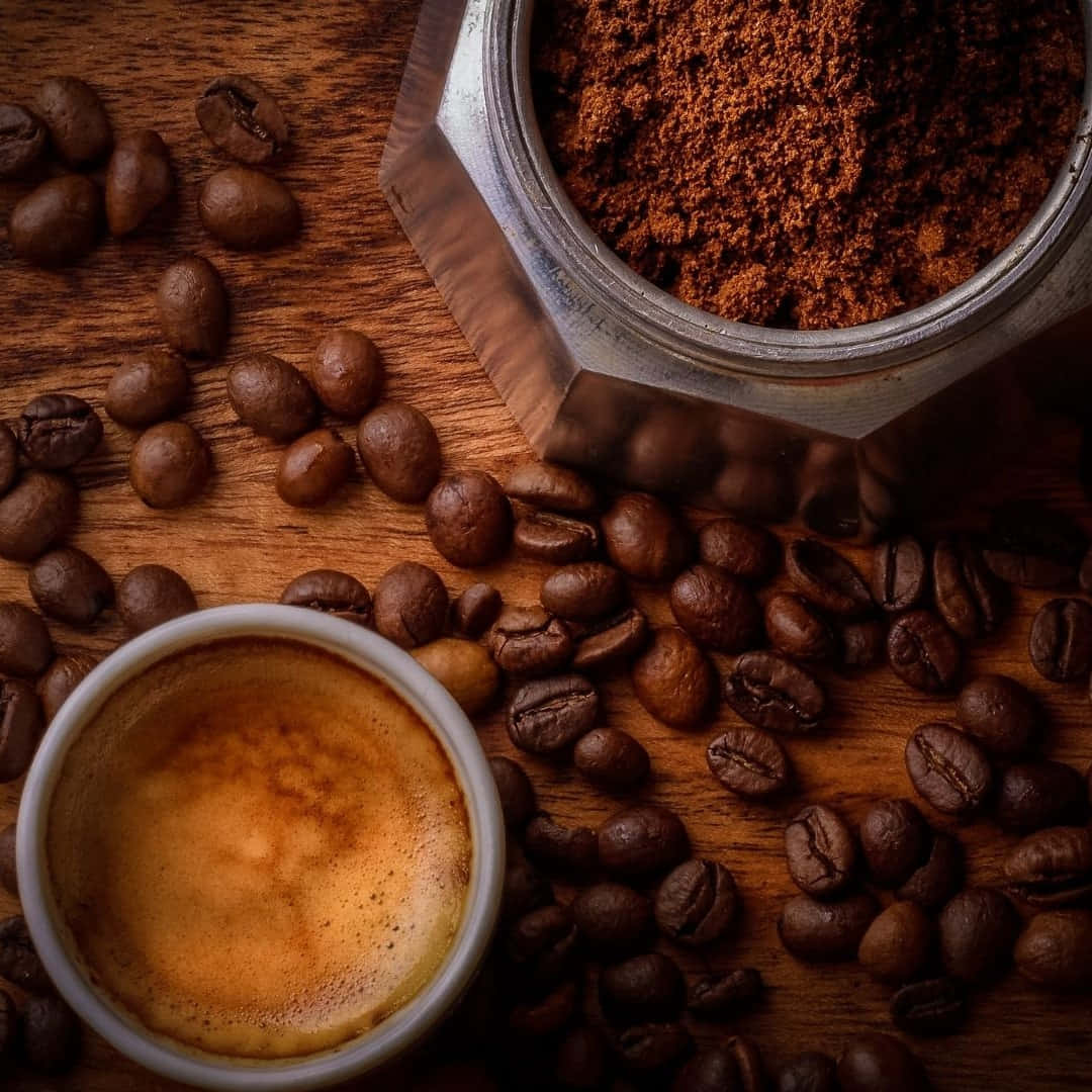 Brown Coffee 桌布 壁紙 — 一杯濃郁的義式濃縮咖啡放在一個六角形研磨機旁邊，研磨機裡裝滿了深色咖啡渣和散落的咖啡豆，置於質樸的木質表面上。