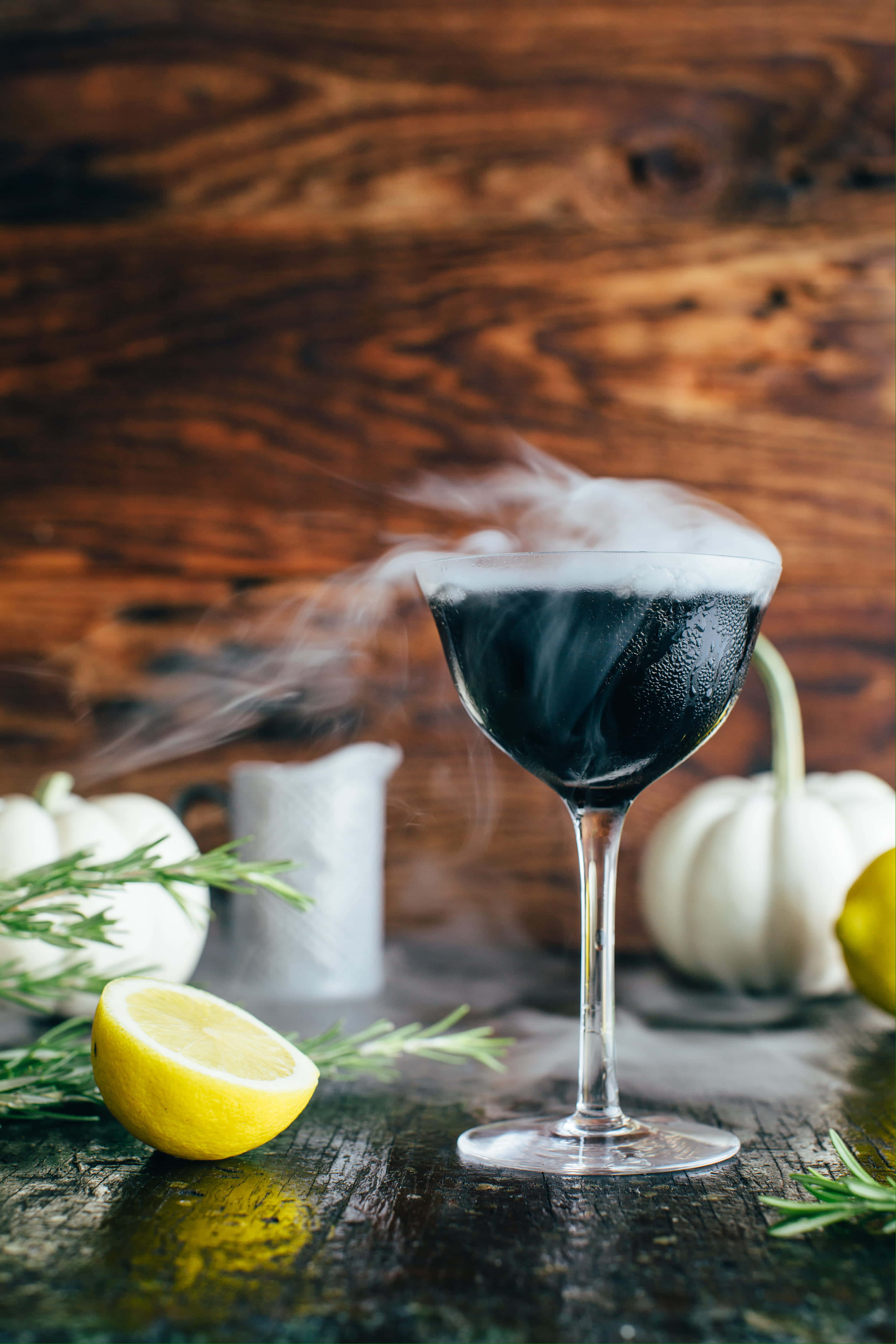 Halloween Drinks 桌布 壁紙 — 一杯煙燻黑色雞尾酒放在高腳杯中，置於鄉村風木桌上，周圍有白色南瓜、檸檬和迷迭香。