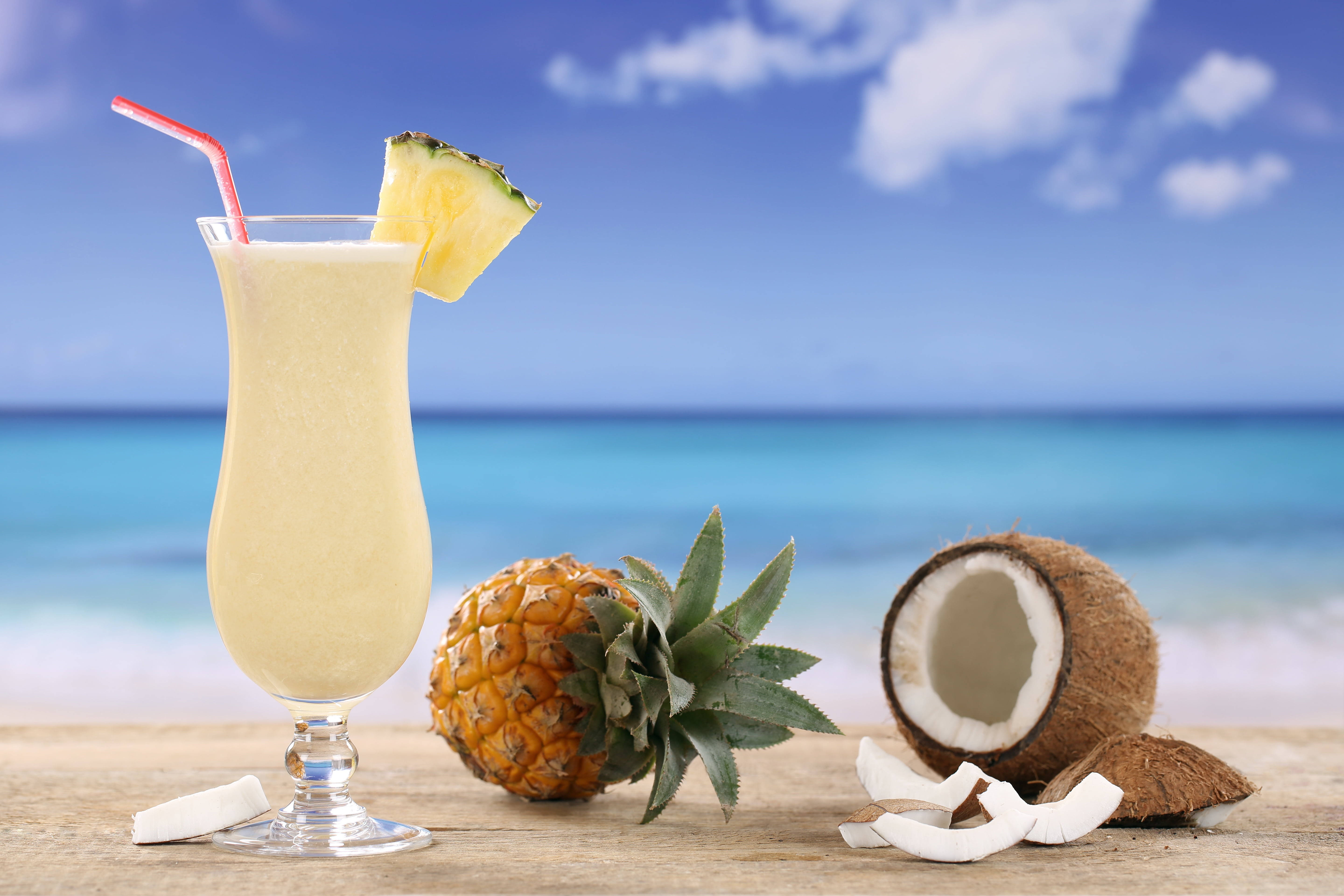 Pineapple 桌布 壁紙 — 一杯熱帶鳳梨飲品，配有紅色吸管和新鮮水果裝飾，放在木桌上，背景是充滿活力的沙灘。
