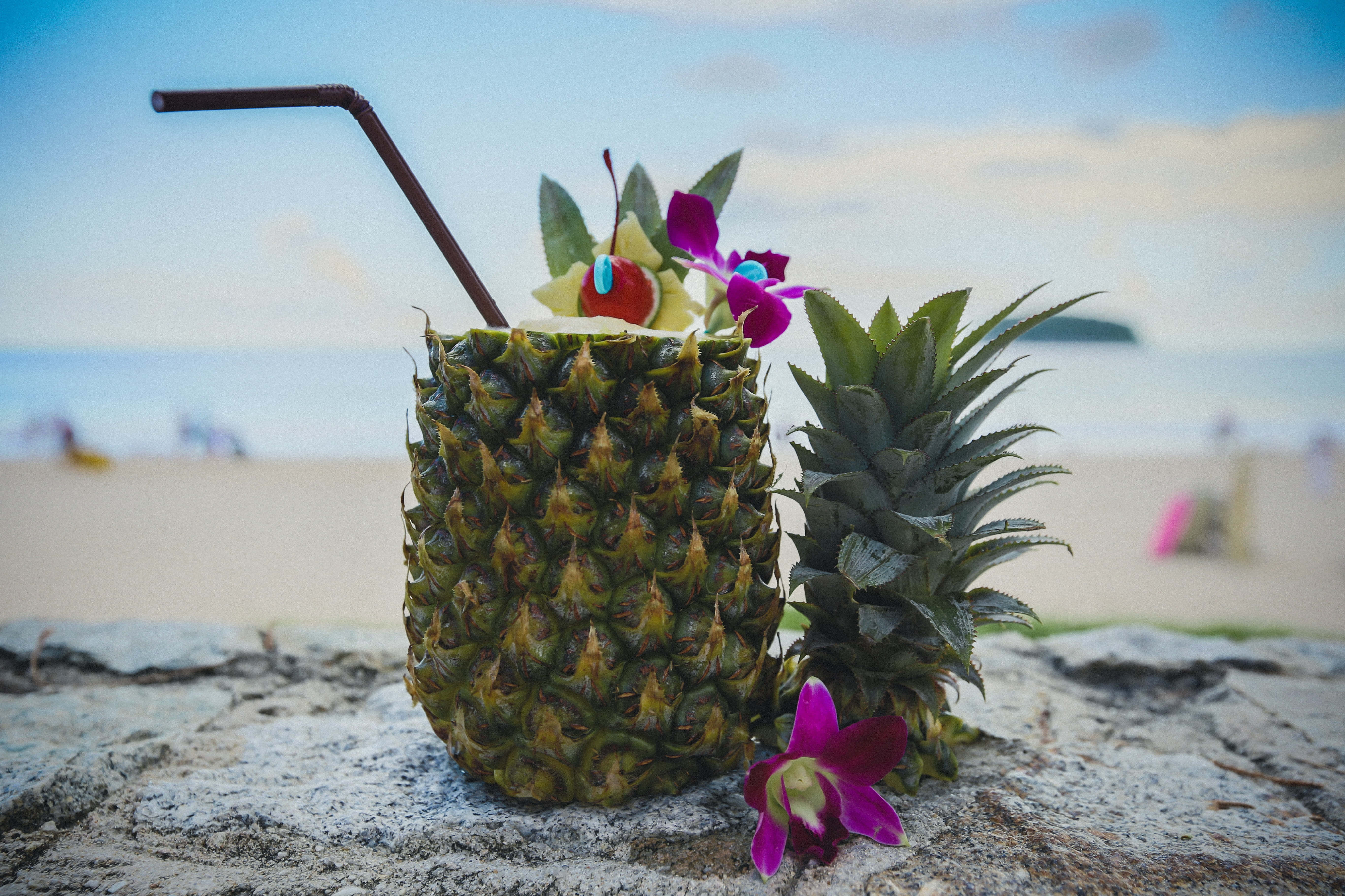 Summer Drinks 桌布 壁紙 — 一杯熱帶鳳梨飲品，飾以花朵和櫻桃，放在陽光沙灘的石頭上，背景是海洋。