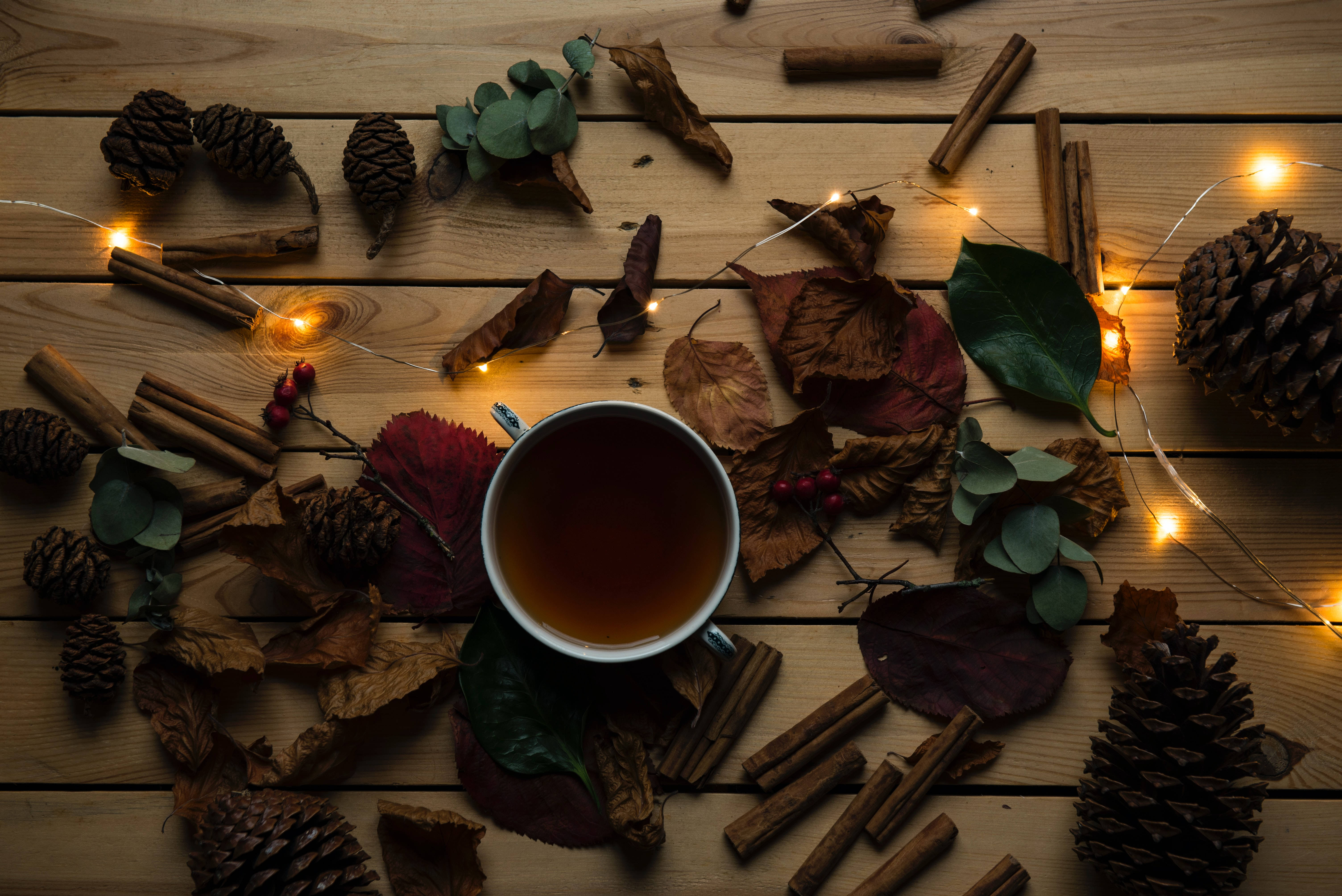 Cozy 桌布 壁紙 — 一杯熱茶放在木桌上，周圍環繞著秋葉、松果、肉桂棒和仙女燈。
