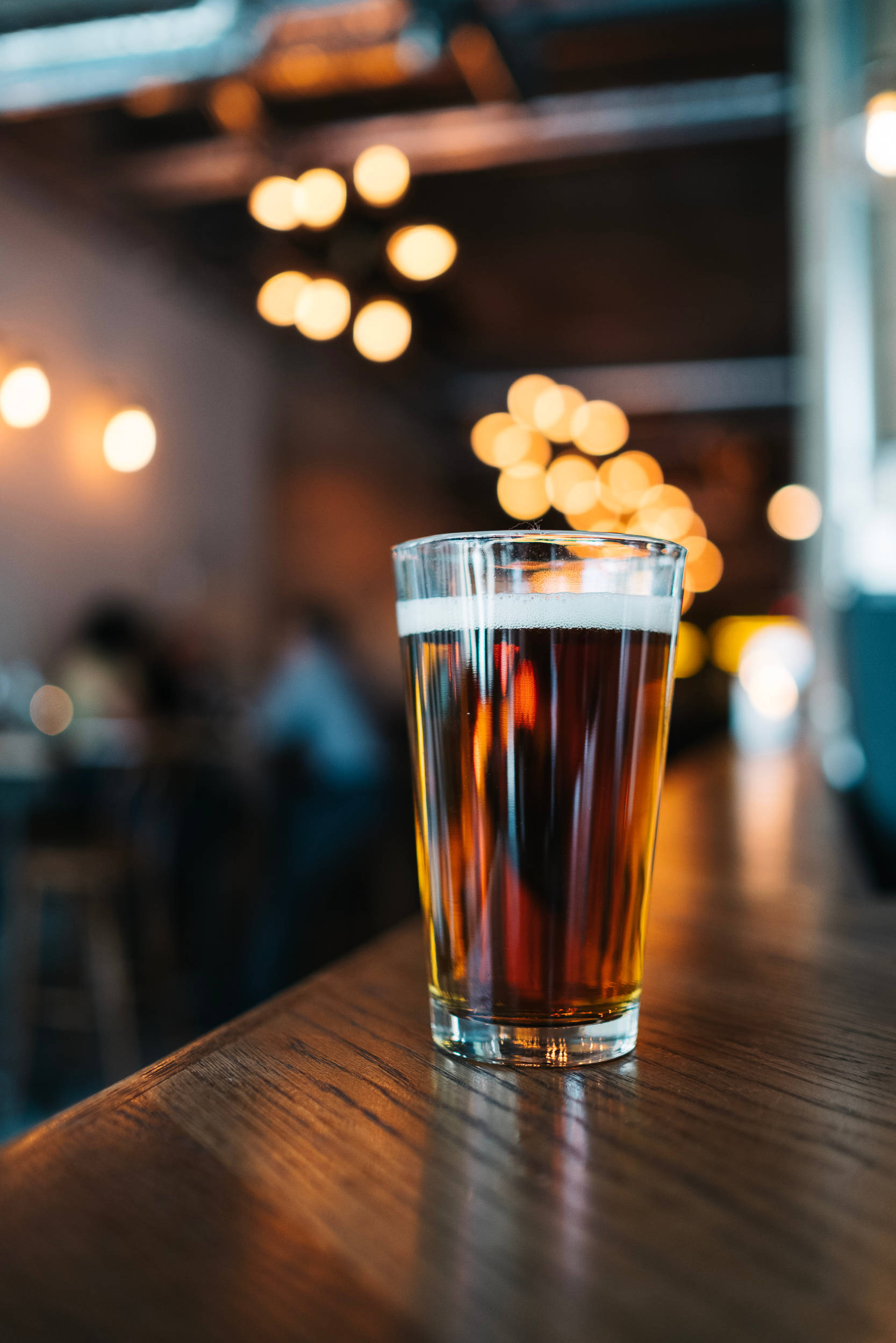 Beer 桌布 壁紙 — 一杯琥珀色啤酒，頂部有泡沫，放在拋光木吧檯上，背景是溫暖的散景燈光。