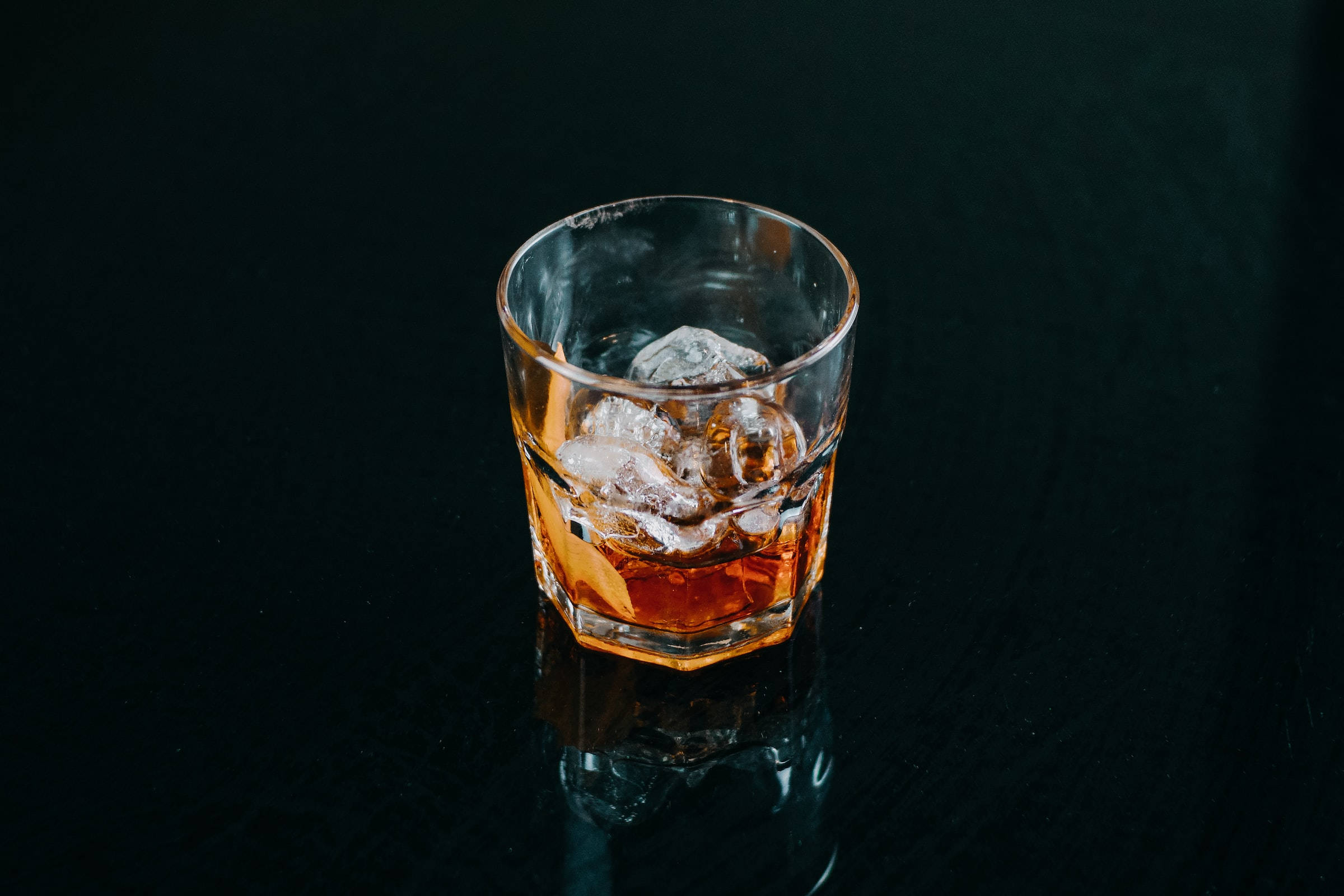 alcohol 桌布 壁紙 — 一杯琥珀色威士忌，帶有正在融化的冰塊，放在光滑的黑色表面上，反射出其溫暖的光芒。