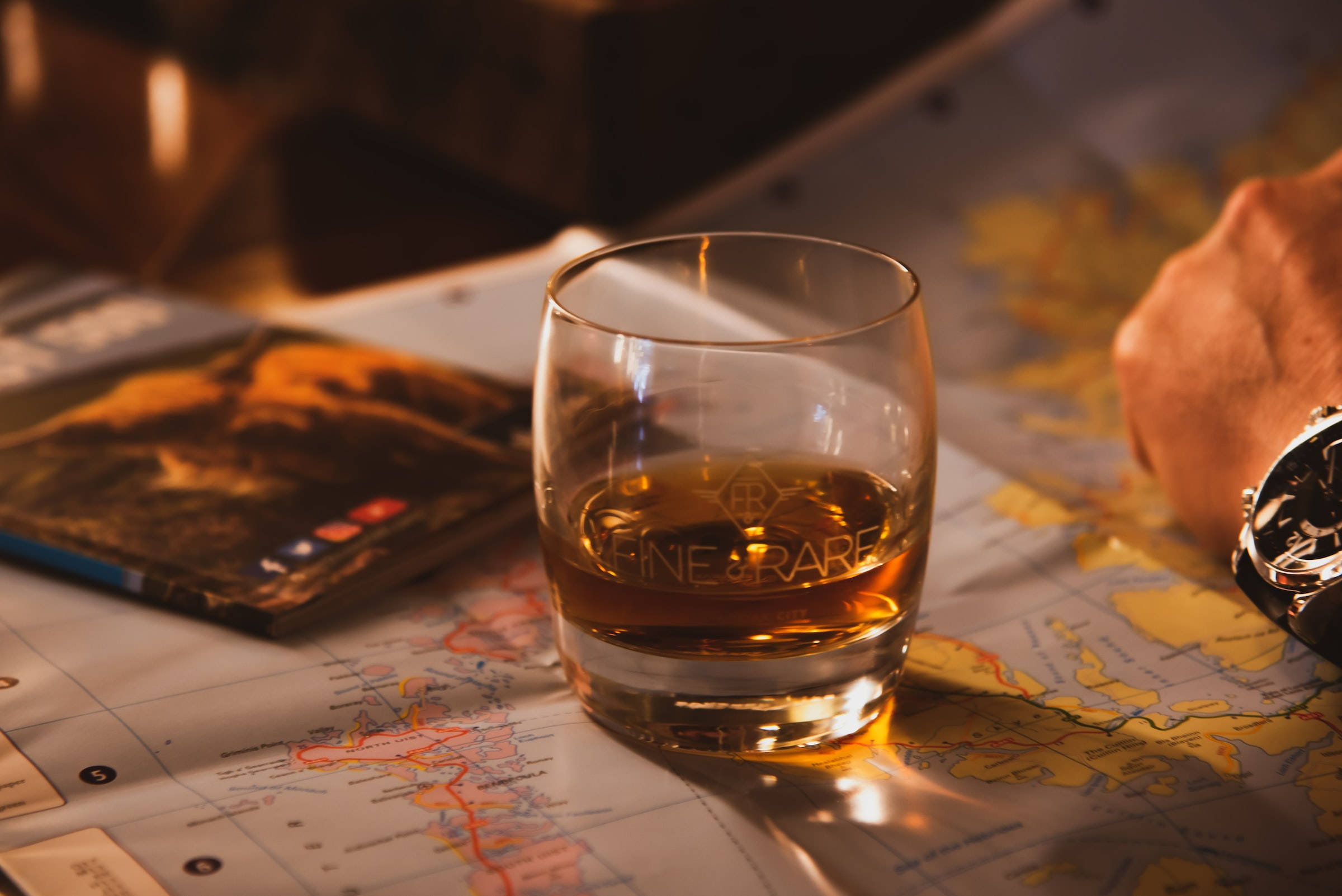Drink 桌布 壁紙 — 一杯琥珀色威士忌放在世界地圖上，旁邊有旅遊指南和手錶，在溫暖的金色燈光下。