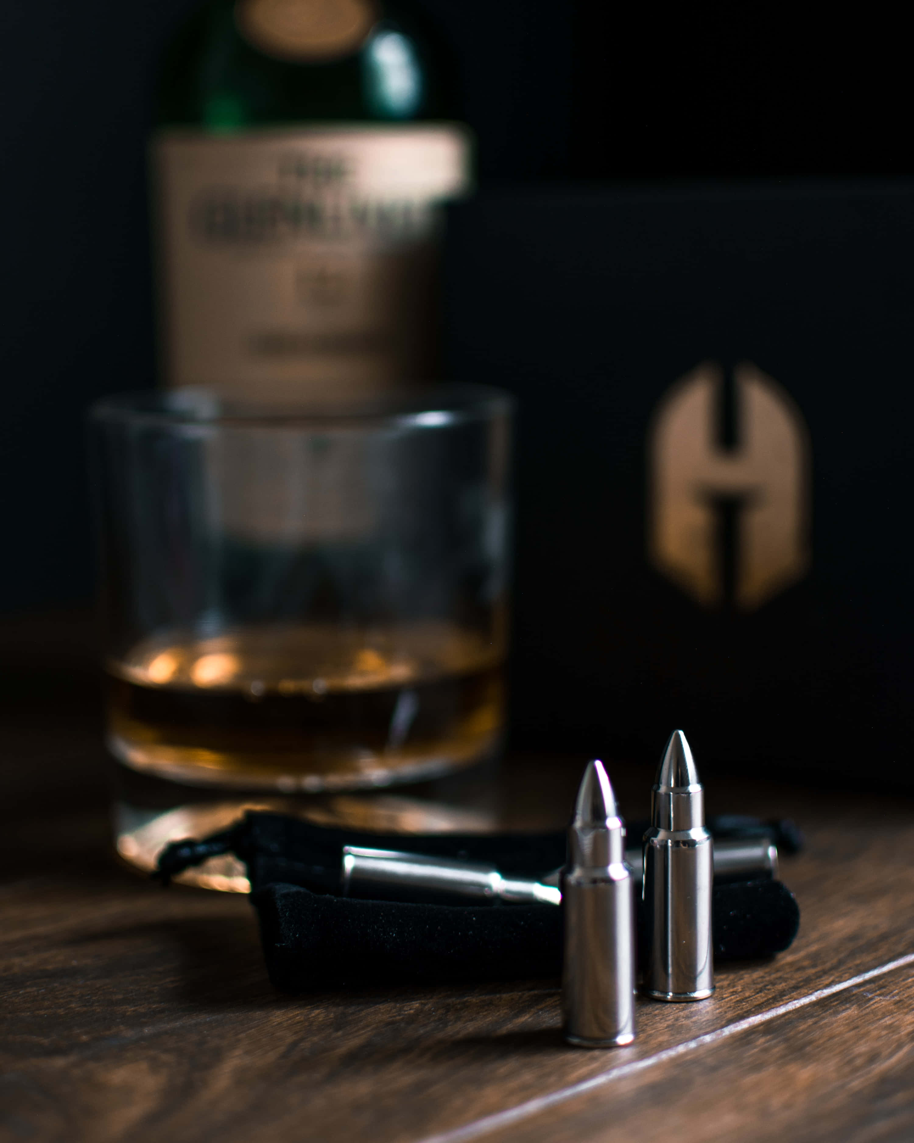 bullet 桌布 壁紙 — 一杯琥珀色威士忌放在木桌上，旁邊有銀色子彈，背景黑暗中有品牌盒子和瓶子。