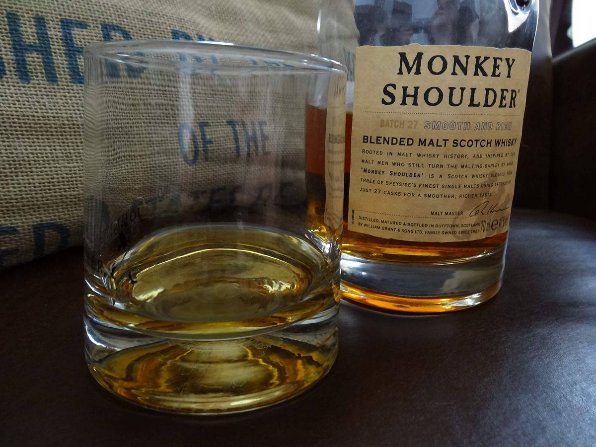 alcohol 桌布 壁紙 — 一杯琥珀色的Monkey Shoulder威士忌放在其酒瓶旁邊，置於深色桌子上，背景是麻布袋。