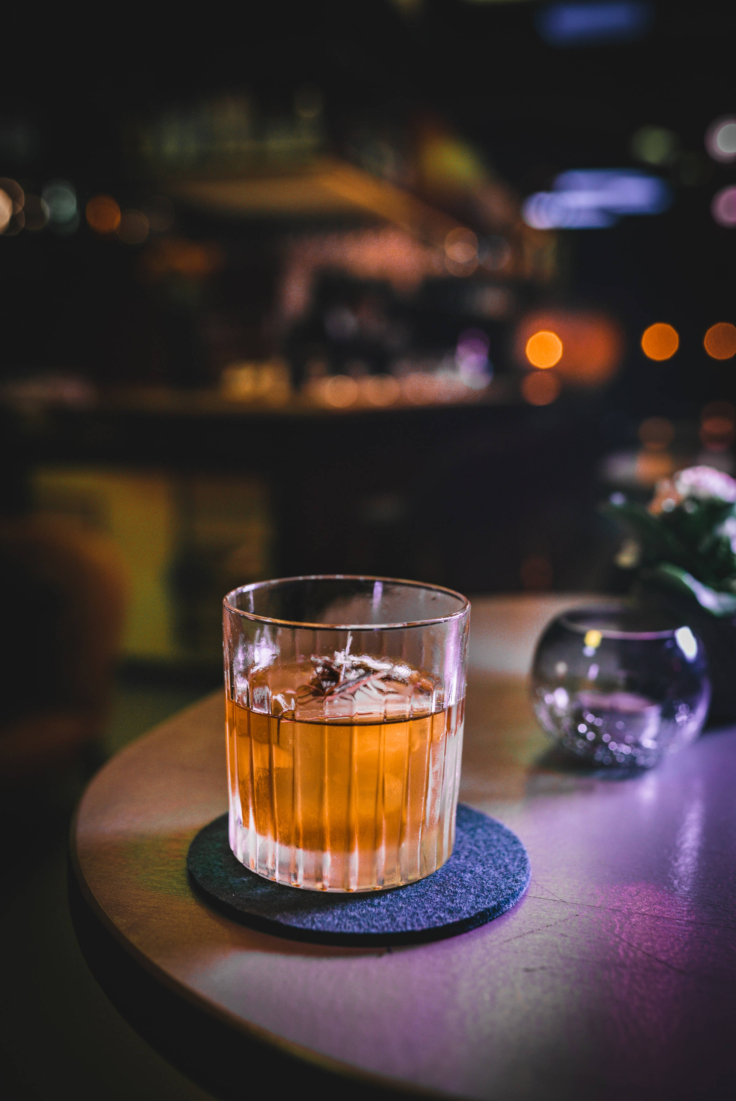 alcohol 桌布 壁紙 — 一杯琥珀色的雞尾酒加冰，盛在帶棱紋的玻璃杯中，放在杯墊上，置於燈光昏暗的酒吧桌上，背景是模糊的城市燈光。