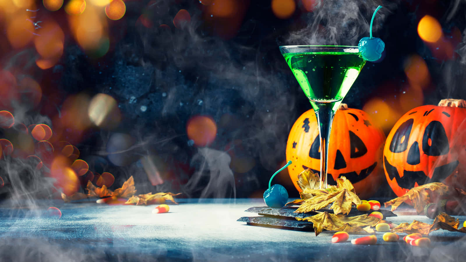 Halloween Cocktails 桌布 壁紙 — 一杯發光綠色萬聖節雞尾酒配藍色櫻桃，旁邊有南瓜燈和糖果玉米，放在煙霧繚繞的桌子上。