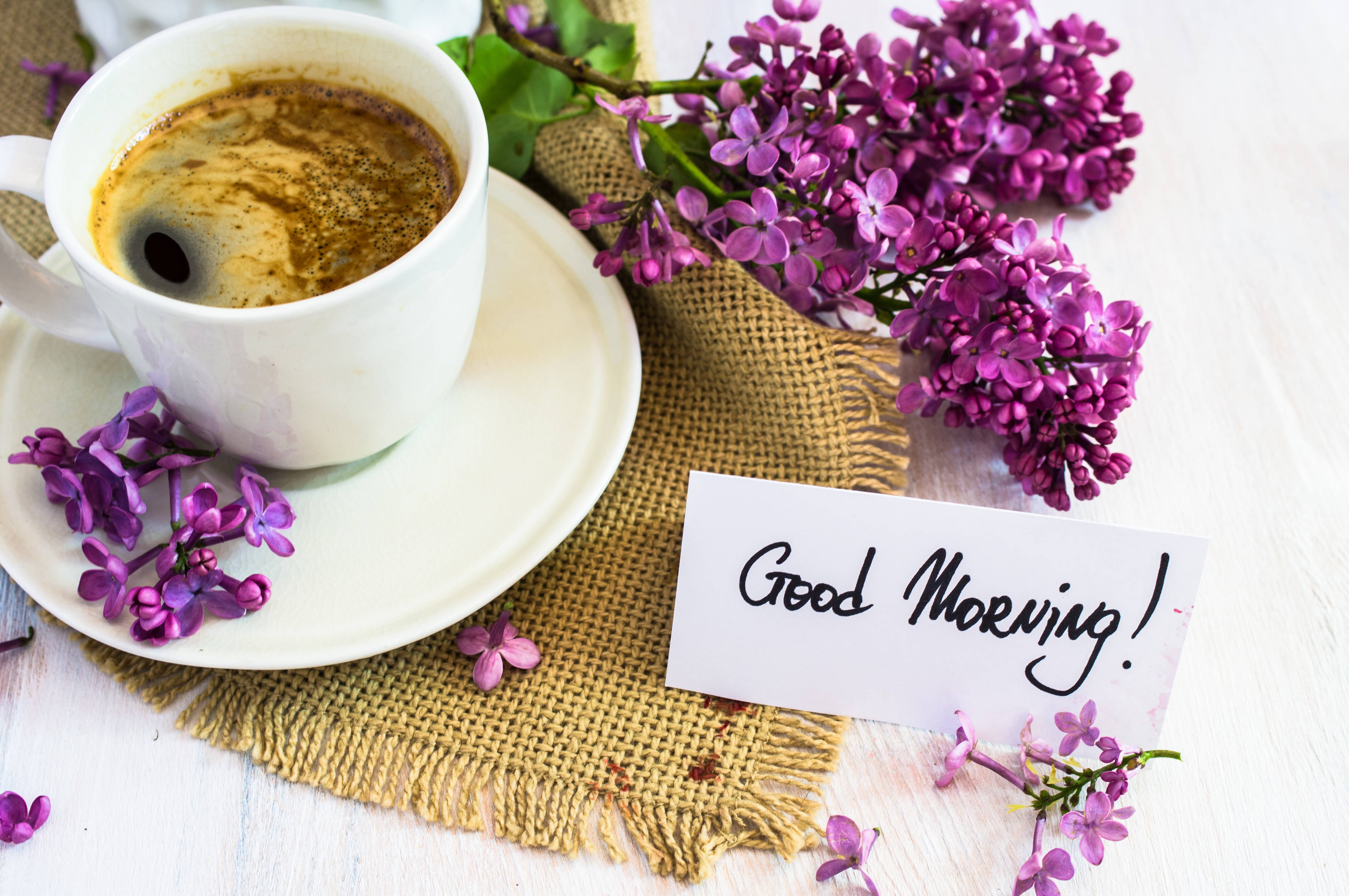 Morning 桌布 壁紙 — 一杯白色咖啡放在麻布墊上，旁邊有紫色丁香花和一張手寫的「早安！」便條。