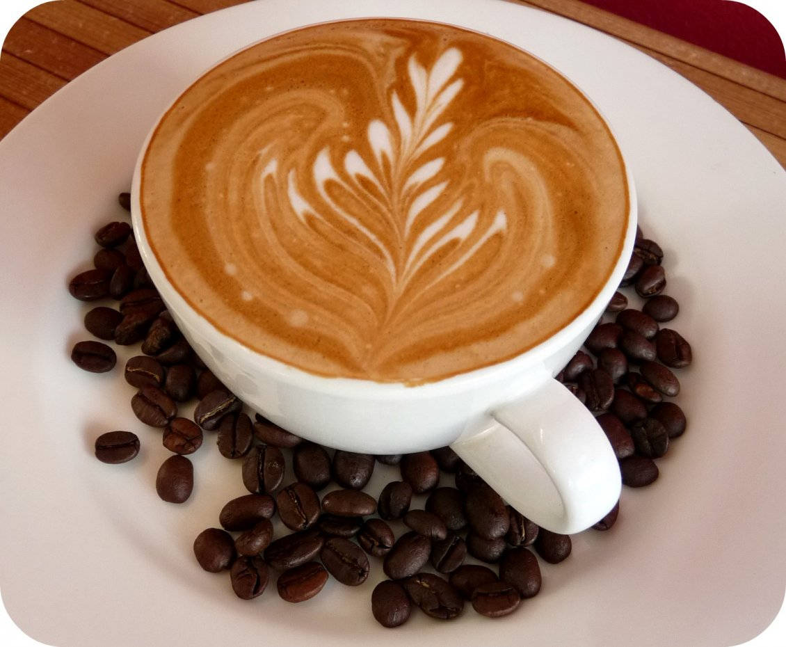 Coffee Aesthetic 桌布 壁紙 — 一杯白色拉花咖啡，上面有葉子圖案，放在盤子上，周圍是烘焙過的咖啡豆。