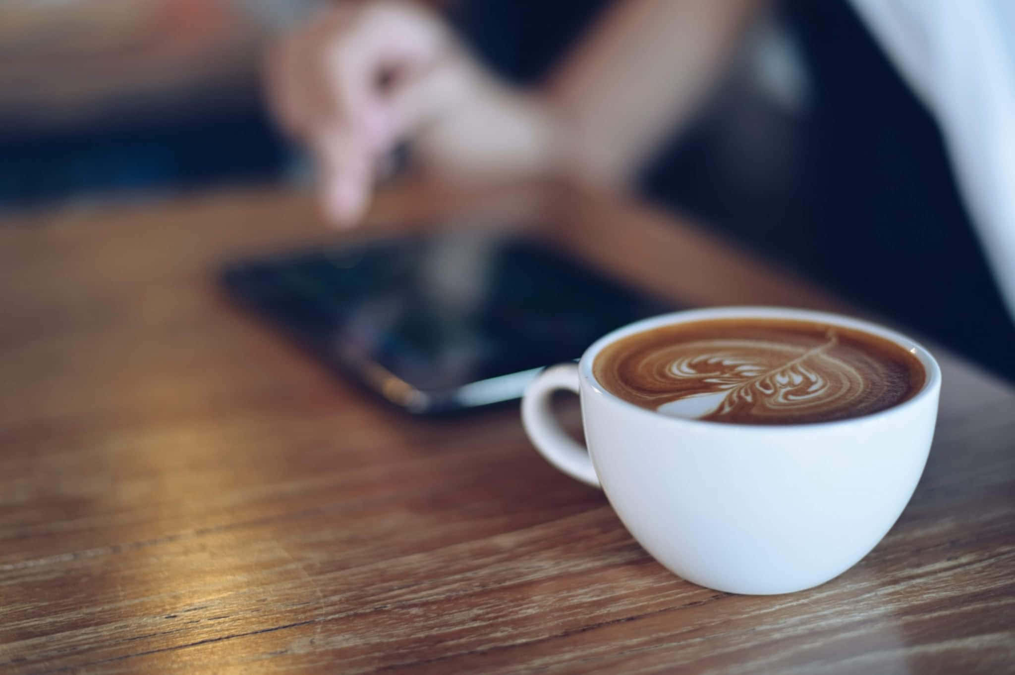 Morning Coffee 桌布 壁紙 — 一杯白色拉花咖啡放在木桌上，背景中一隻模糊的手正在點擊平板電腦。