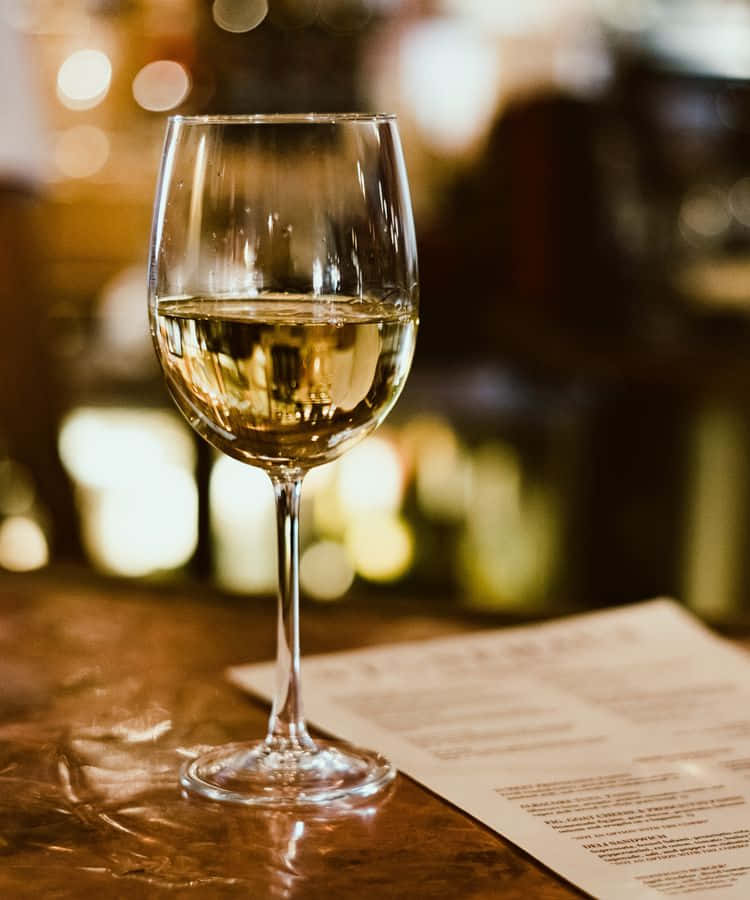 White Wine 桌布 壁紙 — 一杯白酒在木製吧檯上閃耀，背景是模糊溫暖的散景，附近有菜單。