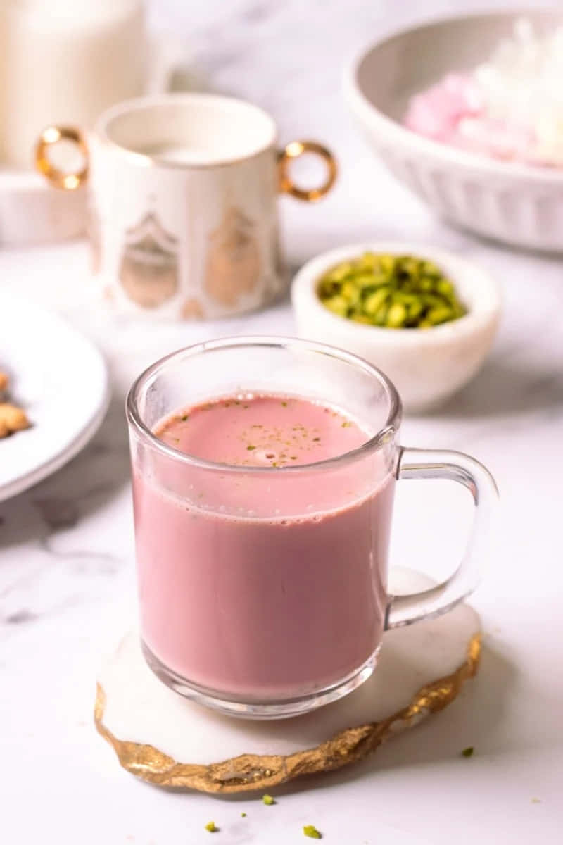 Pink Tea 桌布 壁紙 — 一杯粉紅色牛奶飲品，上面放有開心果，放在金色邊緣的杯墊上，背景有模糊的糕點和碗。