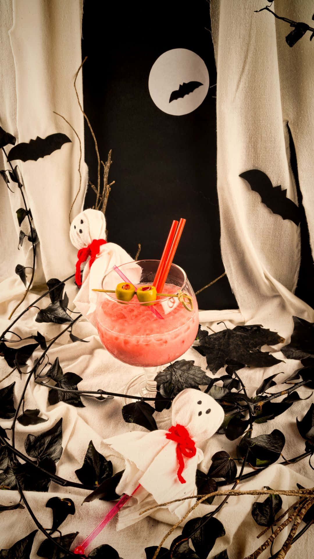 Spooky Cocktails 桌布 壁紙 — 一杯粉紅色萬聖節雞尾酒（配橄欖和吸管）放在蝙蝠月亮背景下的鬼怪裝飾旁。