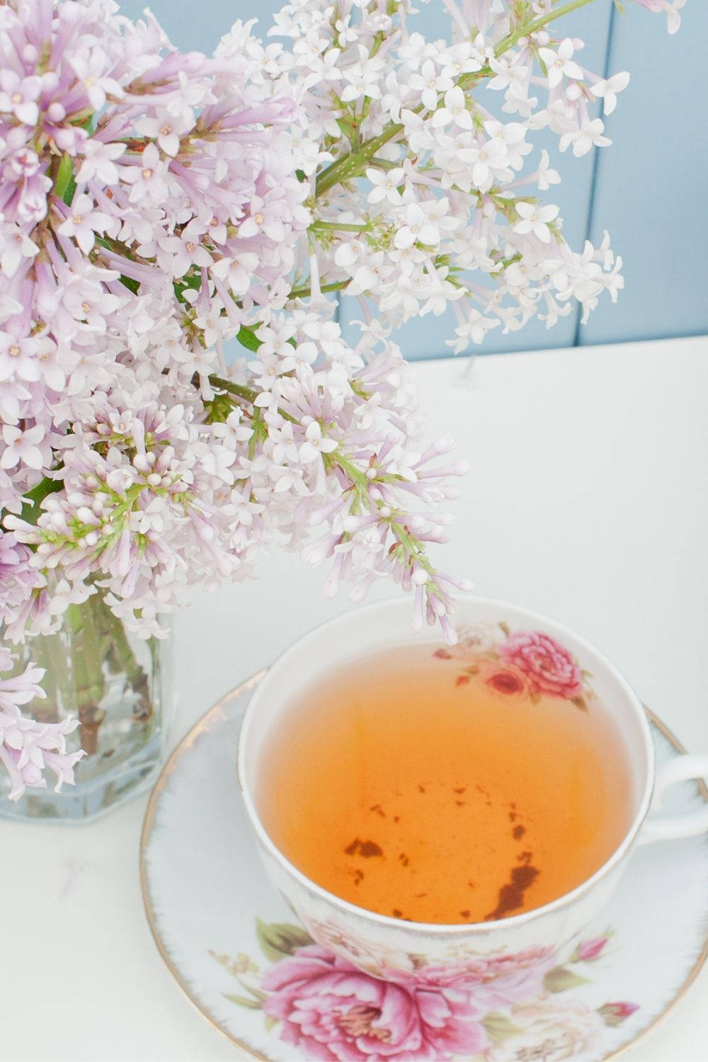 Aesthetic Spring 桌布 壁紙 — 一杯精緻的琥珀色茶放在白色表面上，旁邊是一個插著柔軟粉紅和白色丁香花的花瓶。