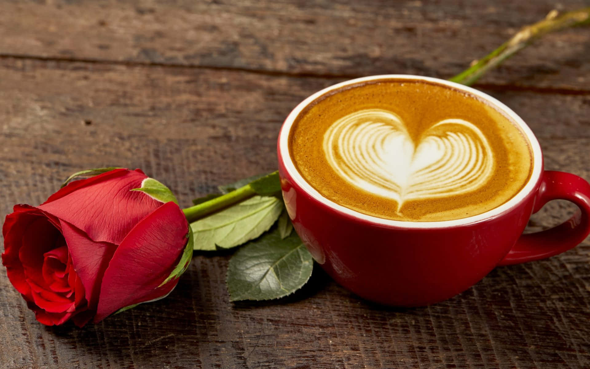 Coffee Aesthetic 桌布 壁紙 — 一杯紅色咖啡杯帶有心形拉花藝術，旁邊放著一朵紅玫瑰，置於鄉村風木桌上。