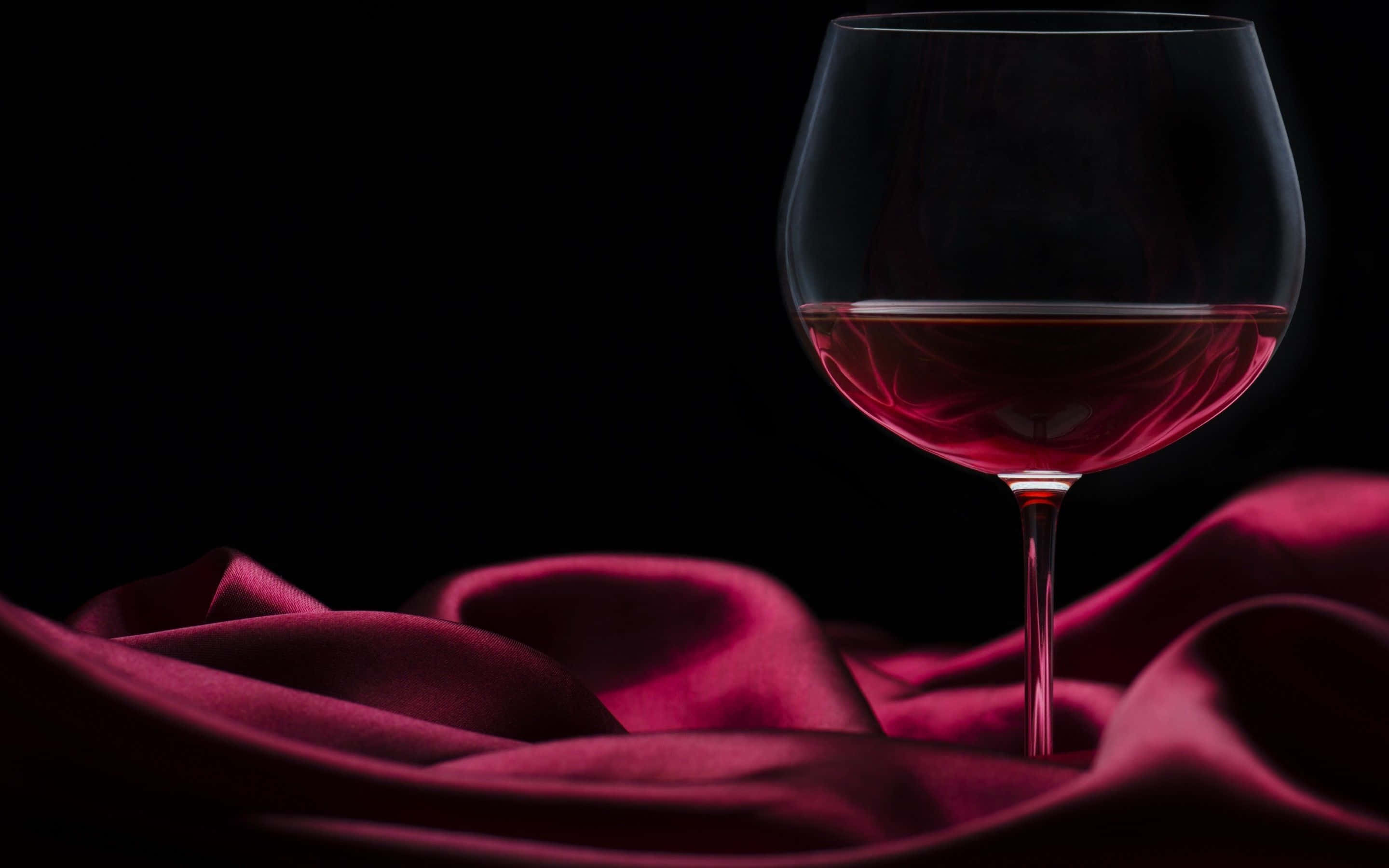 Red Wine Glass 桌布 壁紙 — 一杯紅酒優雅地放置在豐富的酒紅色緞面布料上，背景是純黑色。