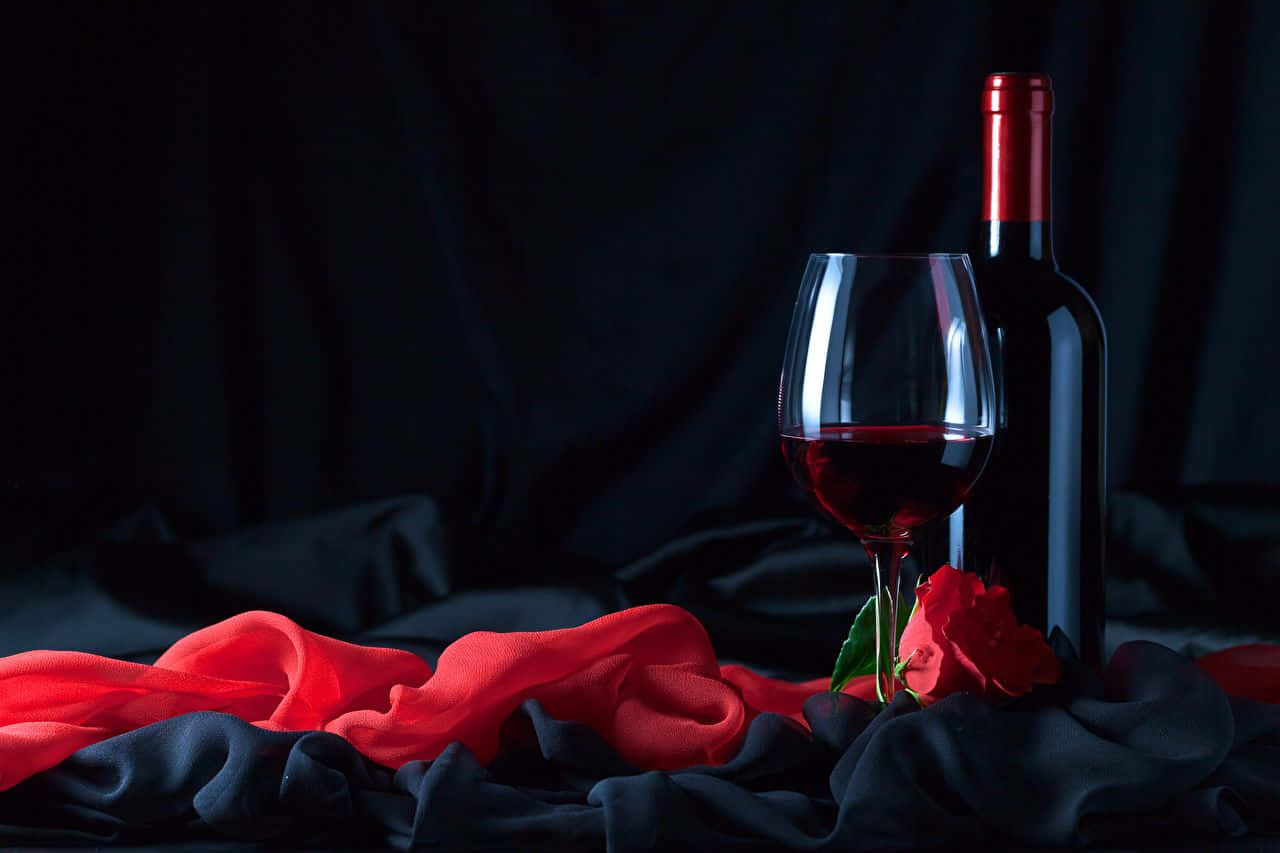 Red Wine Glass 桌布 壁紙 — 一杯紅酒和酒瓶放在深色布料上，旁邊有一朵紅玫瑰和飄逸的紅布，背景為黑色。