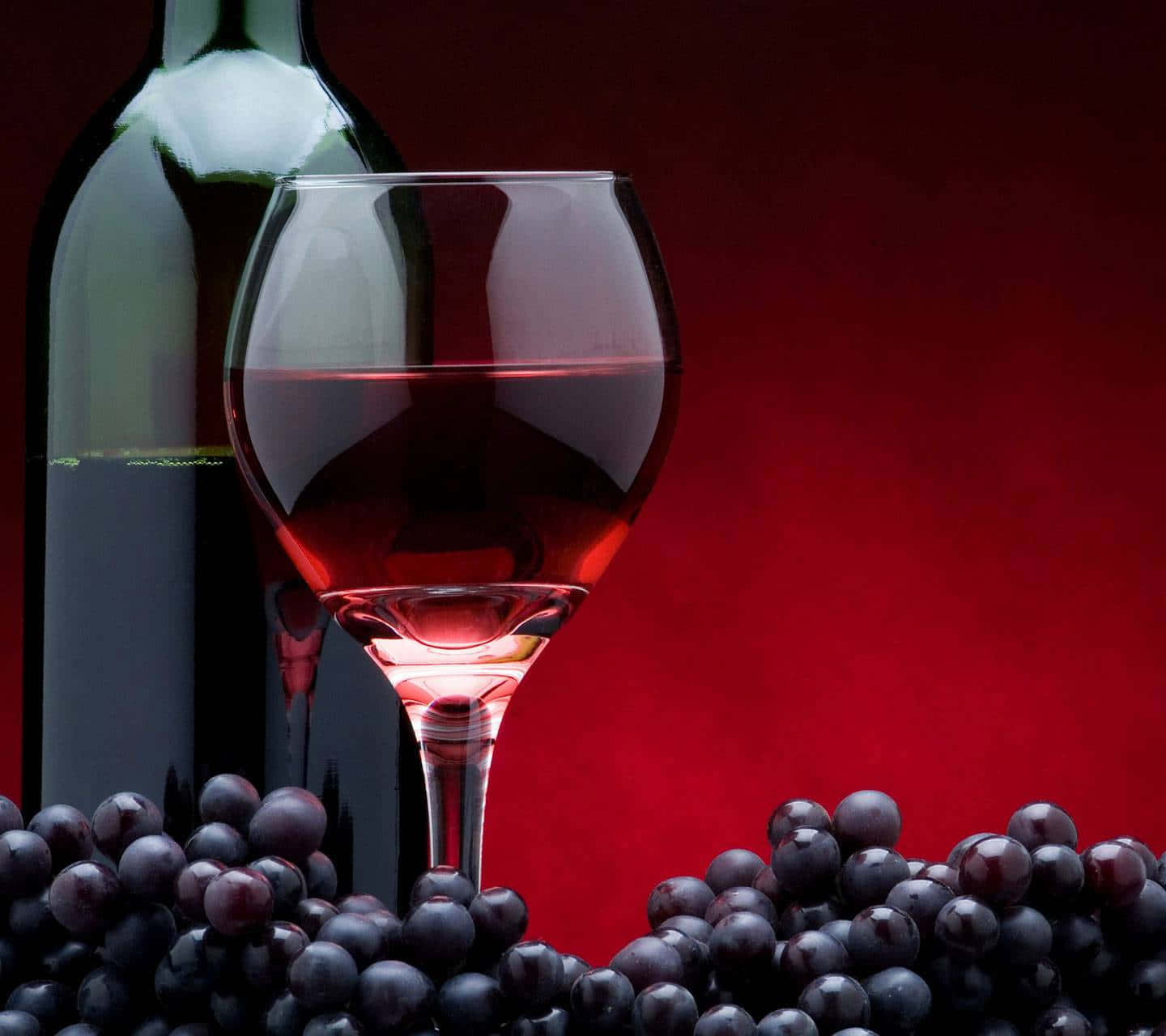 Red Wine Glass 桌布 壁紙 — 一杯紅酒和酒瓶被深色葡萄環繞，襯以濃郁的紅色背景。