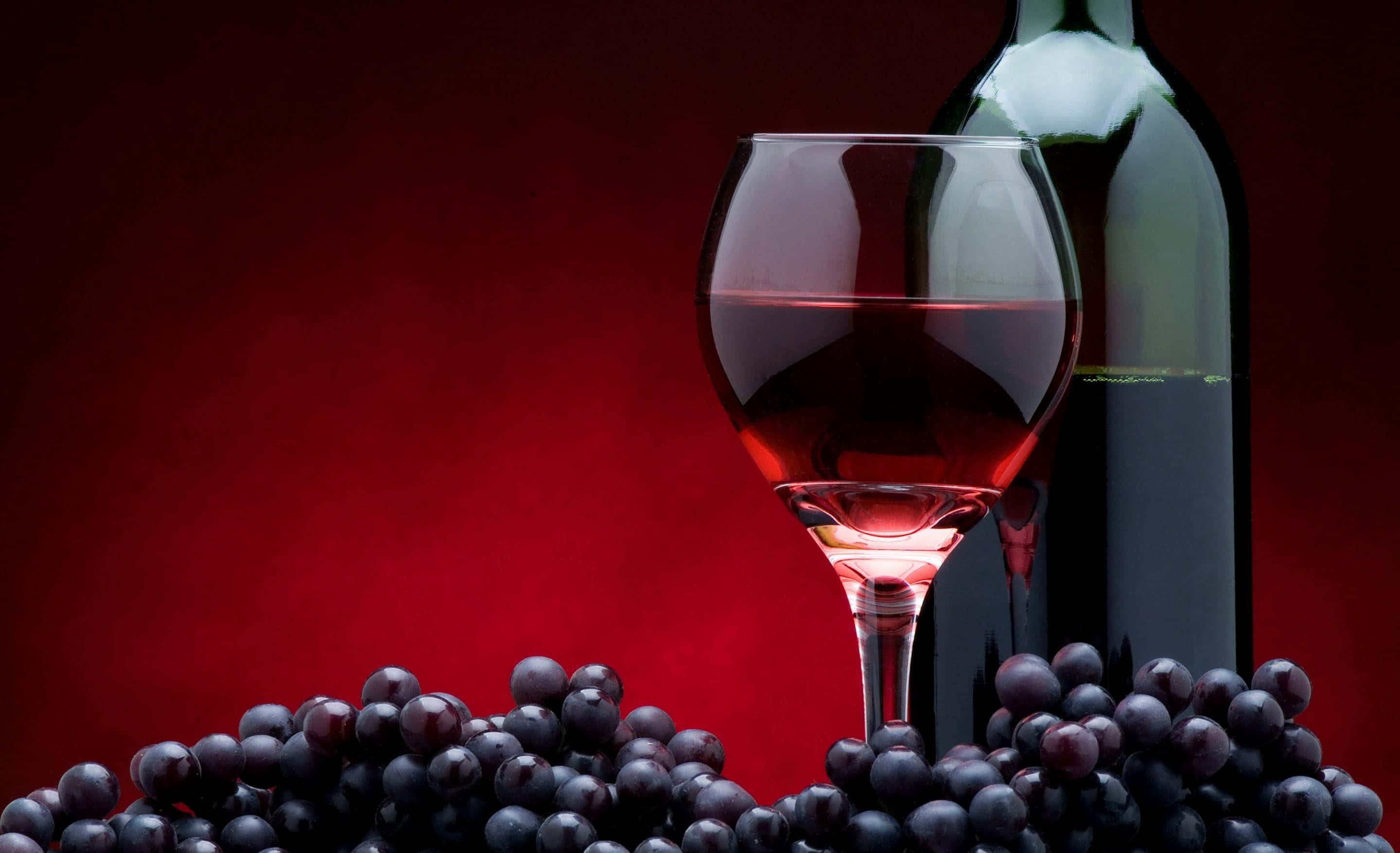 Red Wine Glass 桌布 壁紙 — 一杯紅酒和酒瓶被深色葡萄環繞，襯以濃郁的紅色背景。