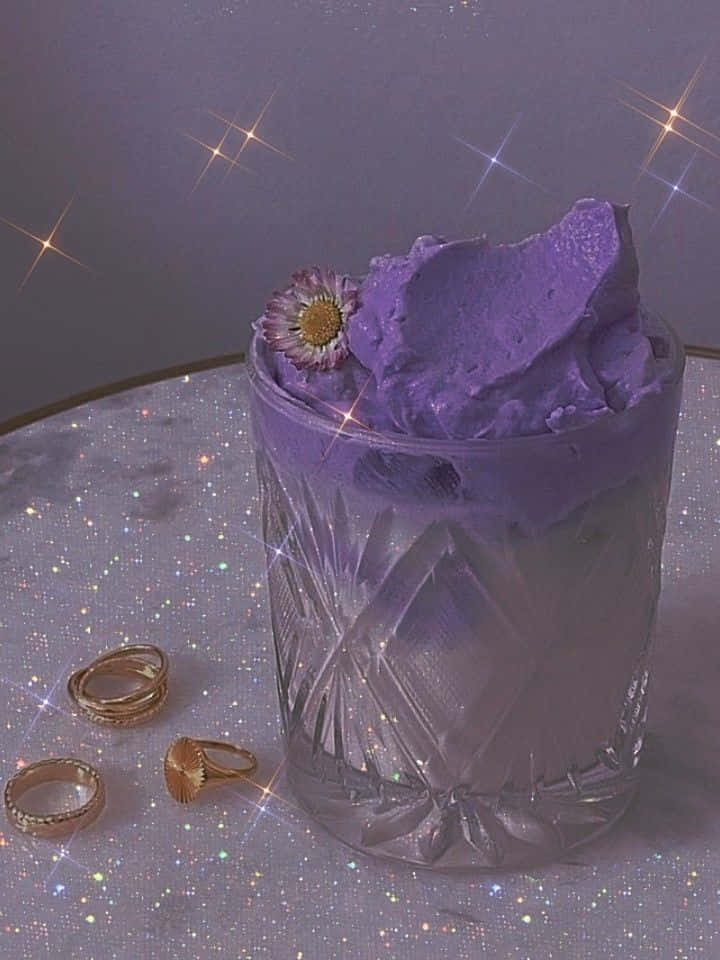 Violet Aesthetic 桌布 壁紙 — 一杯紫色冰淇淋，上面放著一朵花，放在閃閃發光的表面上，附近有閃光和金色戒指。