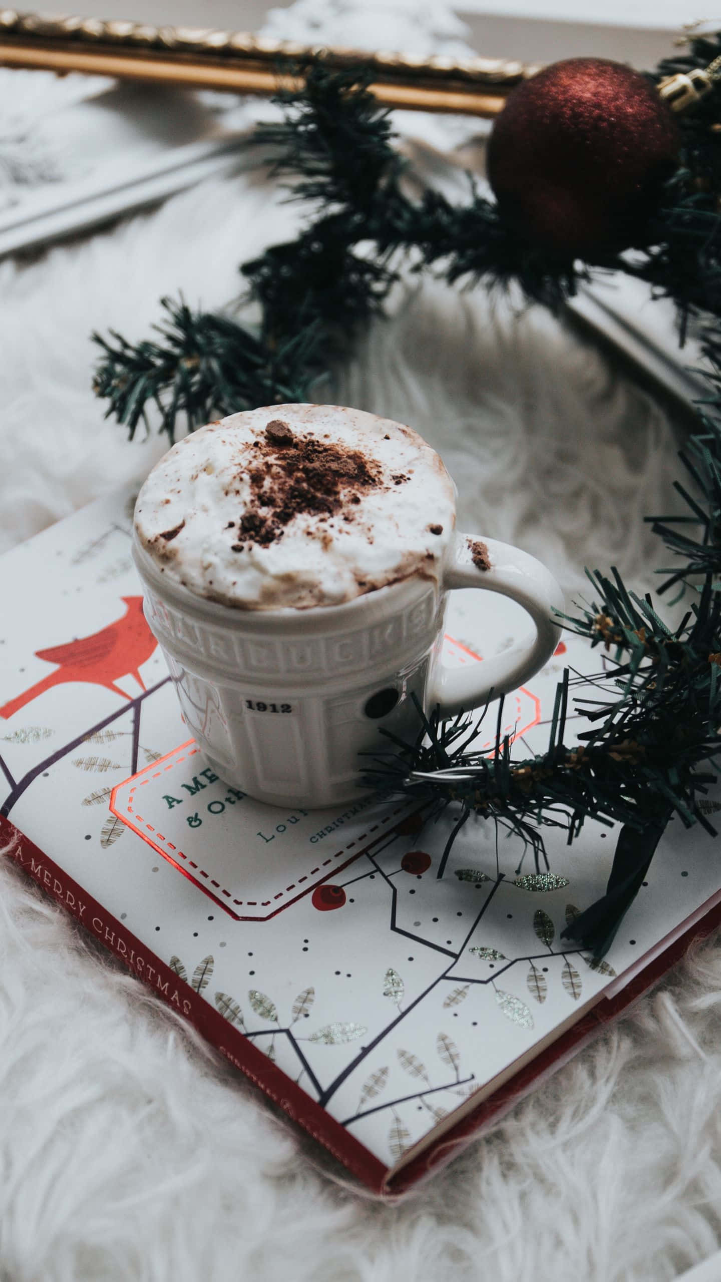Hot Chocolate 桌布 壁紙 — 一杯舒適的節日熱巧克力，配上鮮奶油和可可粉，放在一本節日書上，旁邊有松樹枝和紅色裝飾。