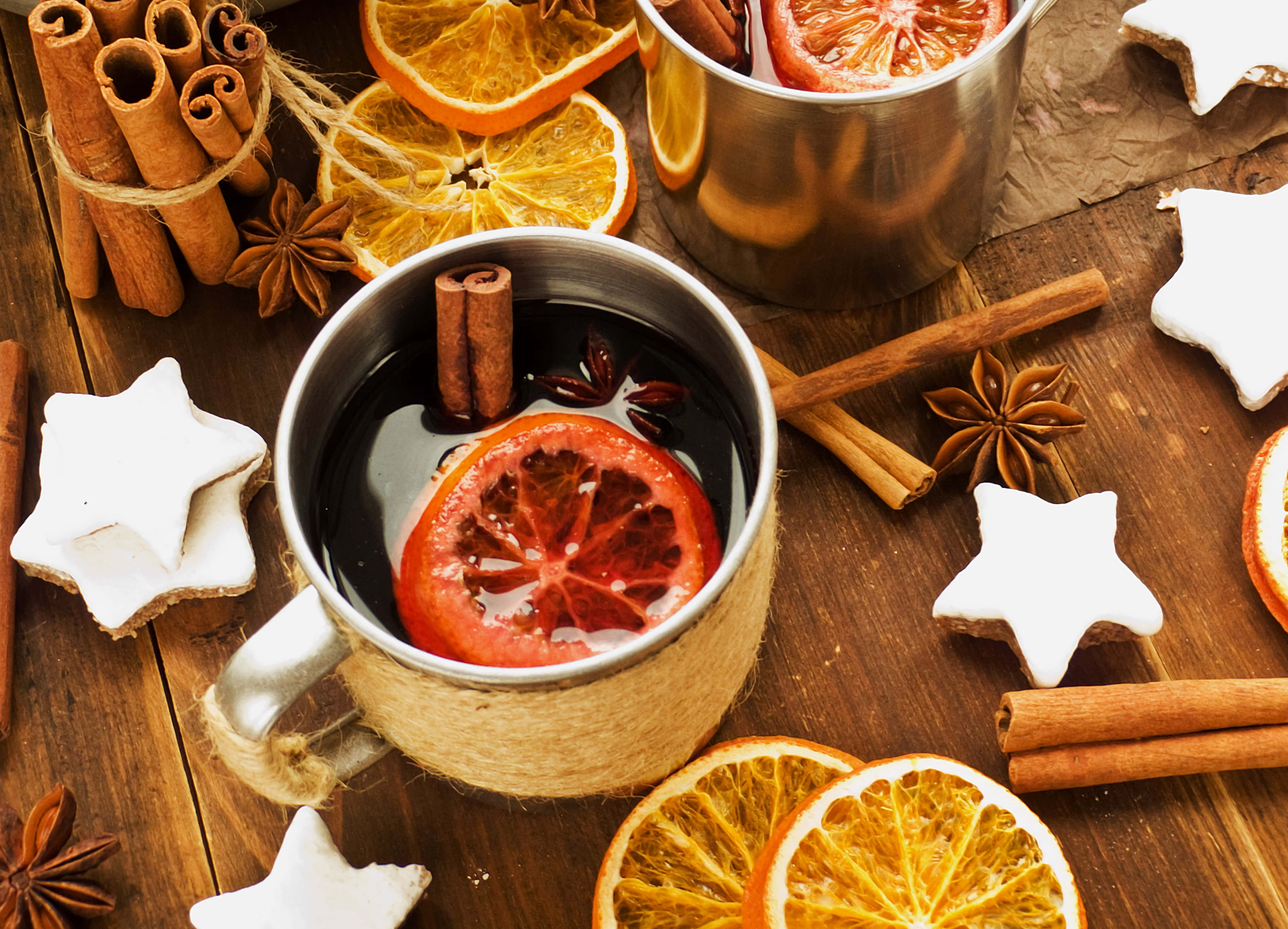 Mulled Wine 桌布 壁紙 — 一杯舒適的香料熱紅酒，配上橙片和肉桂棒，放在質樸的木桌上，周圍環繞著星形餅乾和乾柑橘。