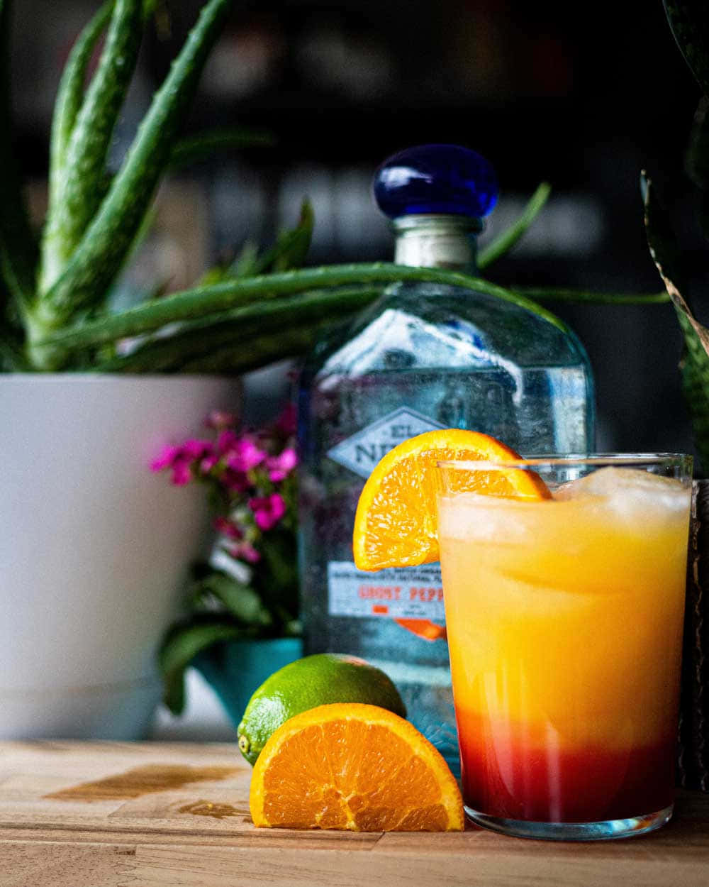 Beautiful Drink 桌布 壁紙 — 一杯色彩鮮豔的漸層雞尾酒，配上冰塊和柳橙片，旁邊放著一瓶琴酒和新鮮柑橘，置於木桌上。