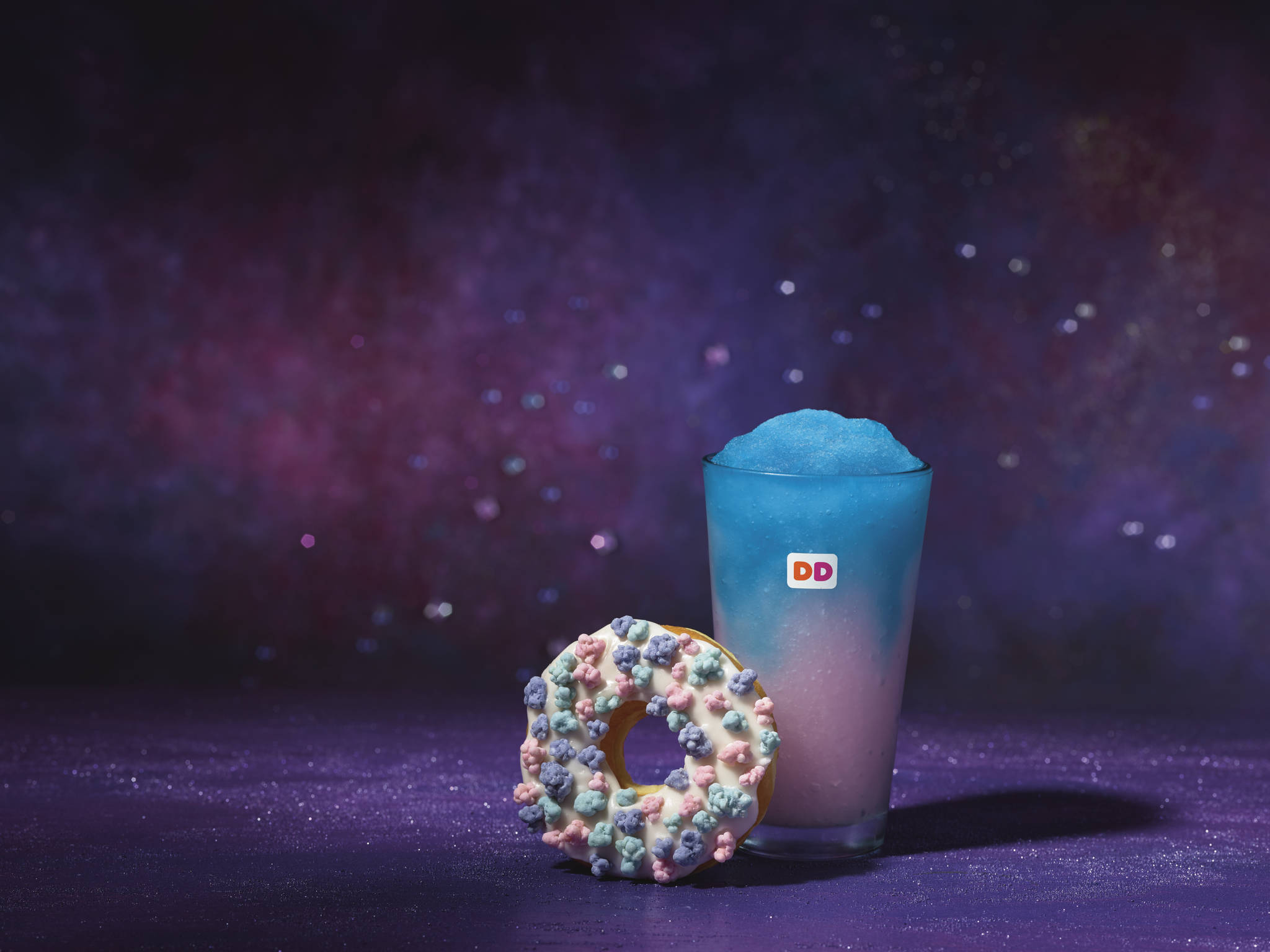 donuts 桌布 壁紙 — 一杯藍色思樂冰飲料和一個灑上糖粒的甜甜圈，放在閃閃發光的紫色表面上，背景是宇宙星空。