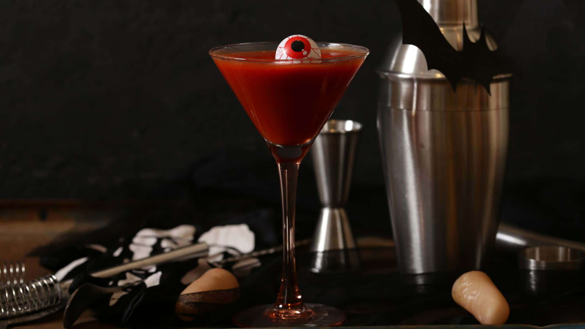 Spooky Cocktails 桌布 壁紙 — 一杯血紅色馬丁尼雞尾酒，上面有恐怖的眼球裝飾，周圍環繞著萬聖節主題的酒吧工具。